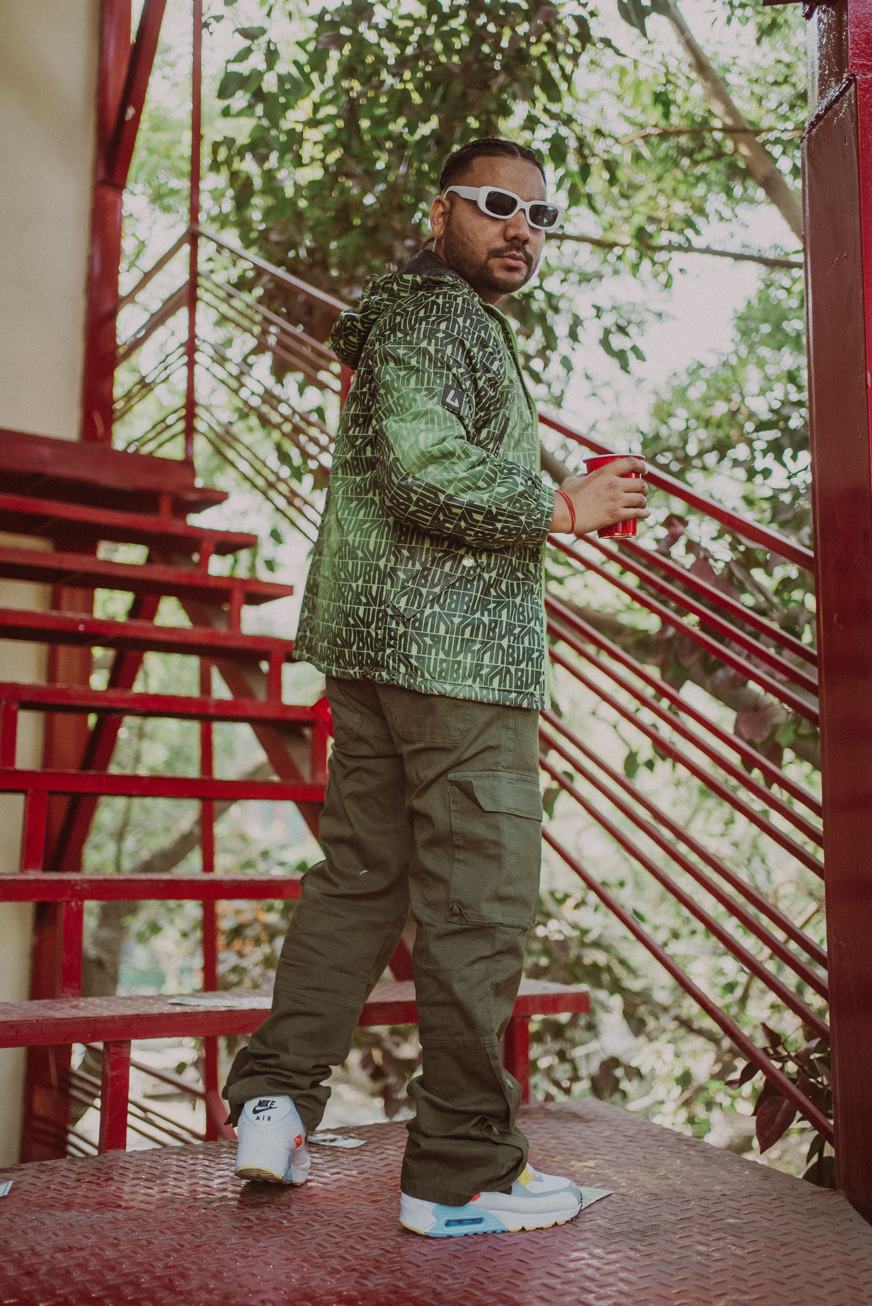 Stylish man in green jacket rockin' sunglasses standing on vibrant red outdoor stairs.