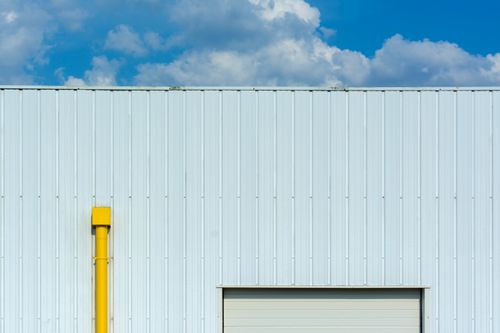 Free A minimalist view of a white industrial building facade with a prominent yellow exhaust pipe against a blue sky with clouds. Stock Photo