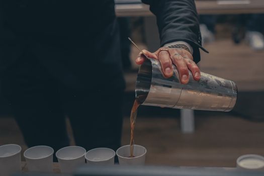 A bartender pours a drink from a shaker into multiple cups during an indoor event.