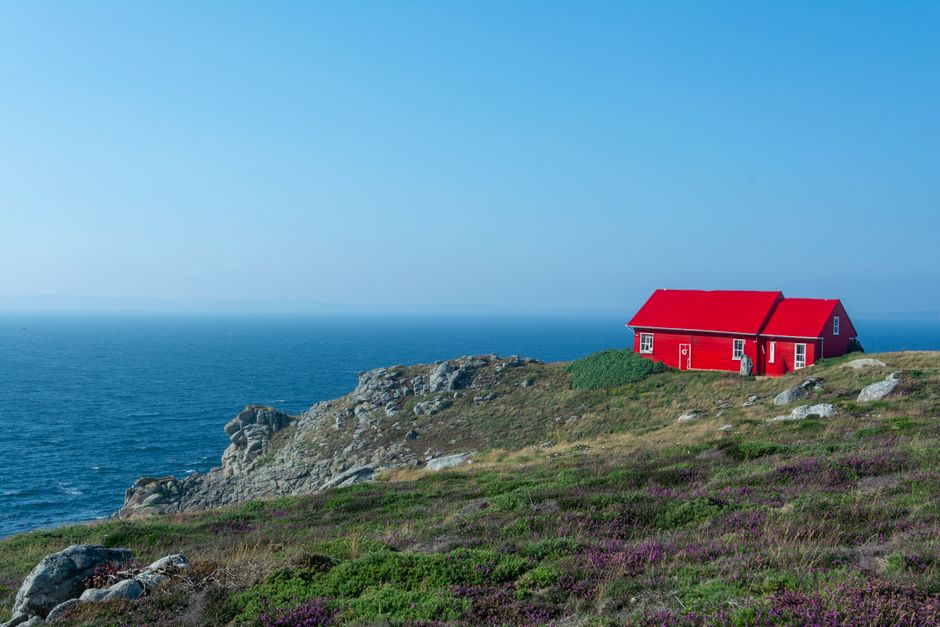 Idyllic red house perched on a rocky coastline under a clear blue sky, overlooking the ocean.