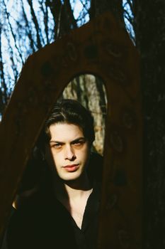 A young man stands outdoors with his reflection captured in an ornate mirror, sunlit against a natural backdrop.