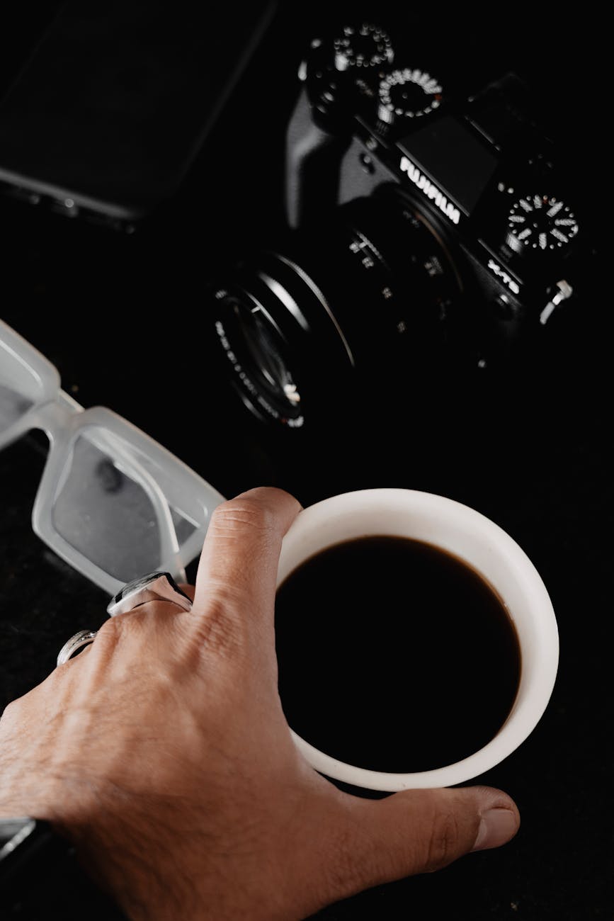 Close-up view of a hand holding a coffee cup next to a camera and glasses on a dark background.