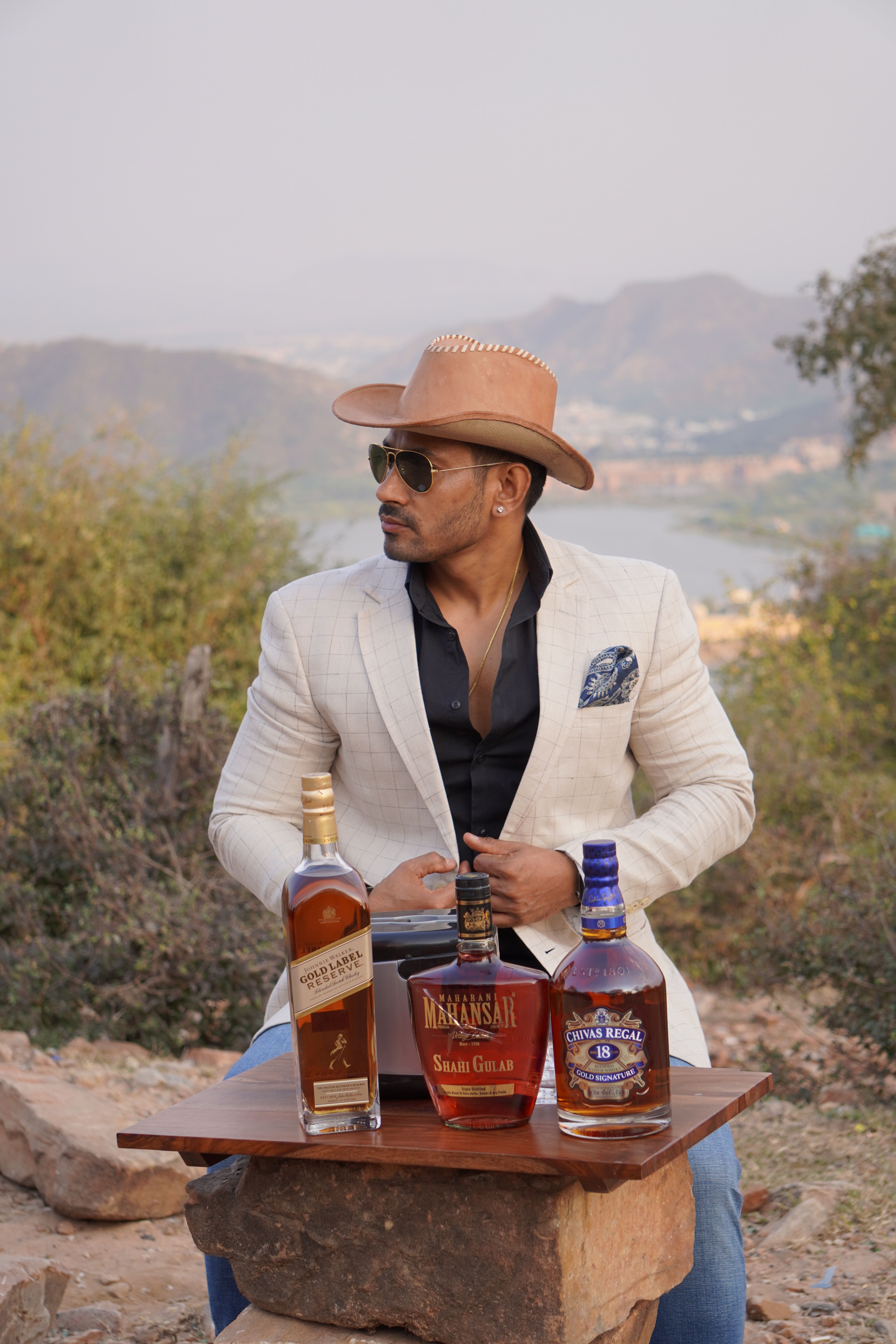 Man in cowboy hat with whisky bottles sitting outdoors with scenic mountain background.