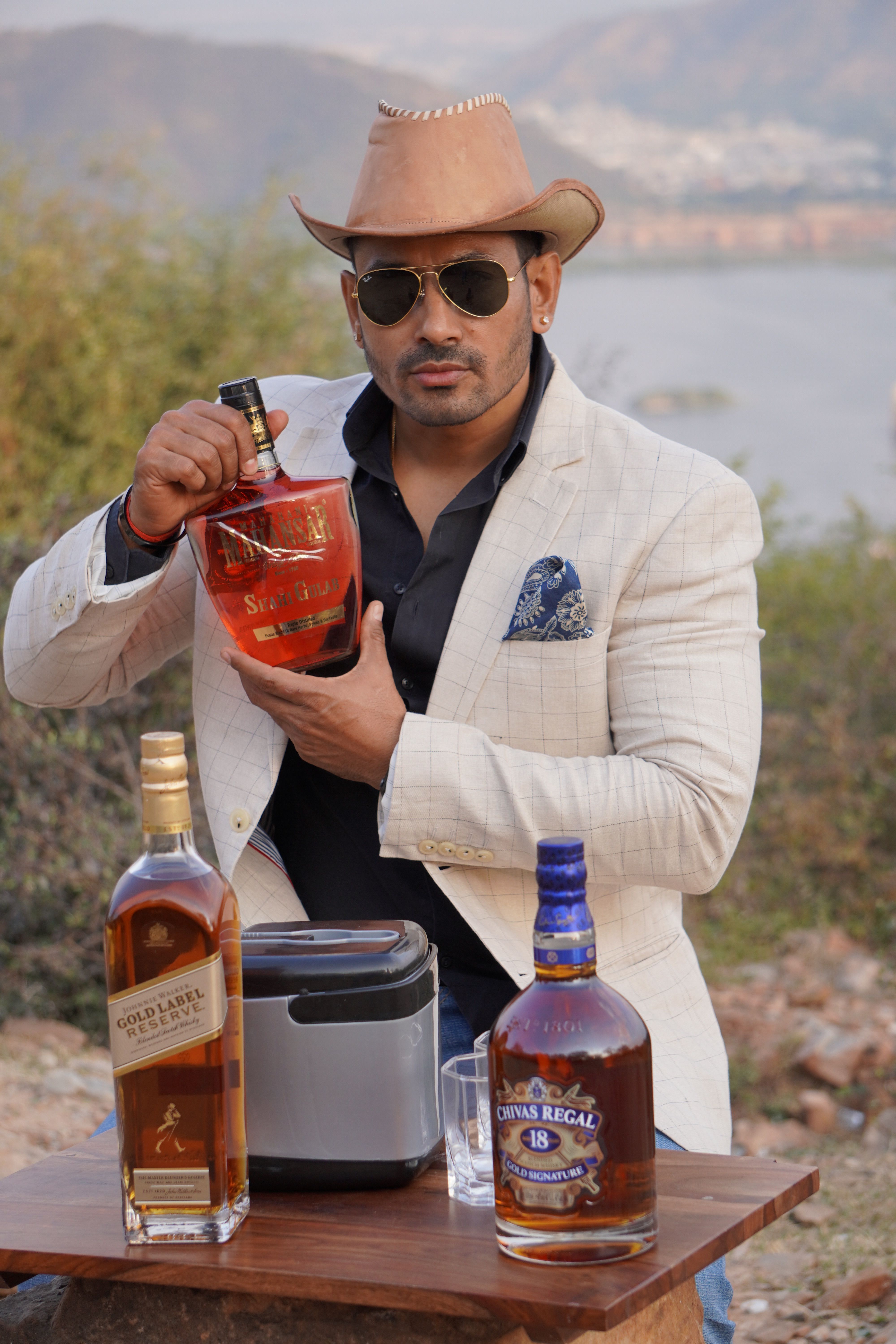 Man in cowboy hat showcasing whiskey bottles on a table outdoors. Elegant and stylish.