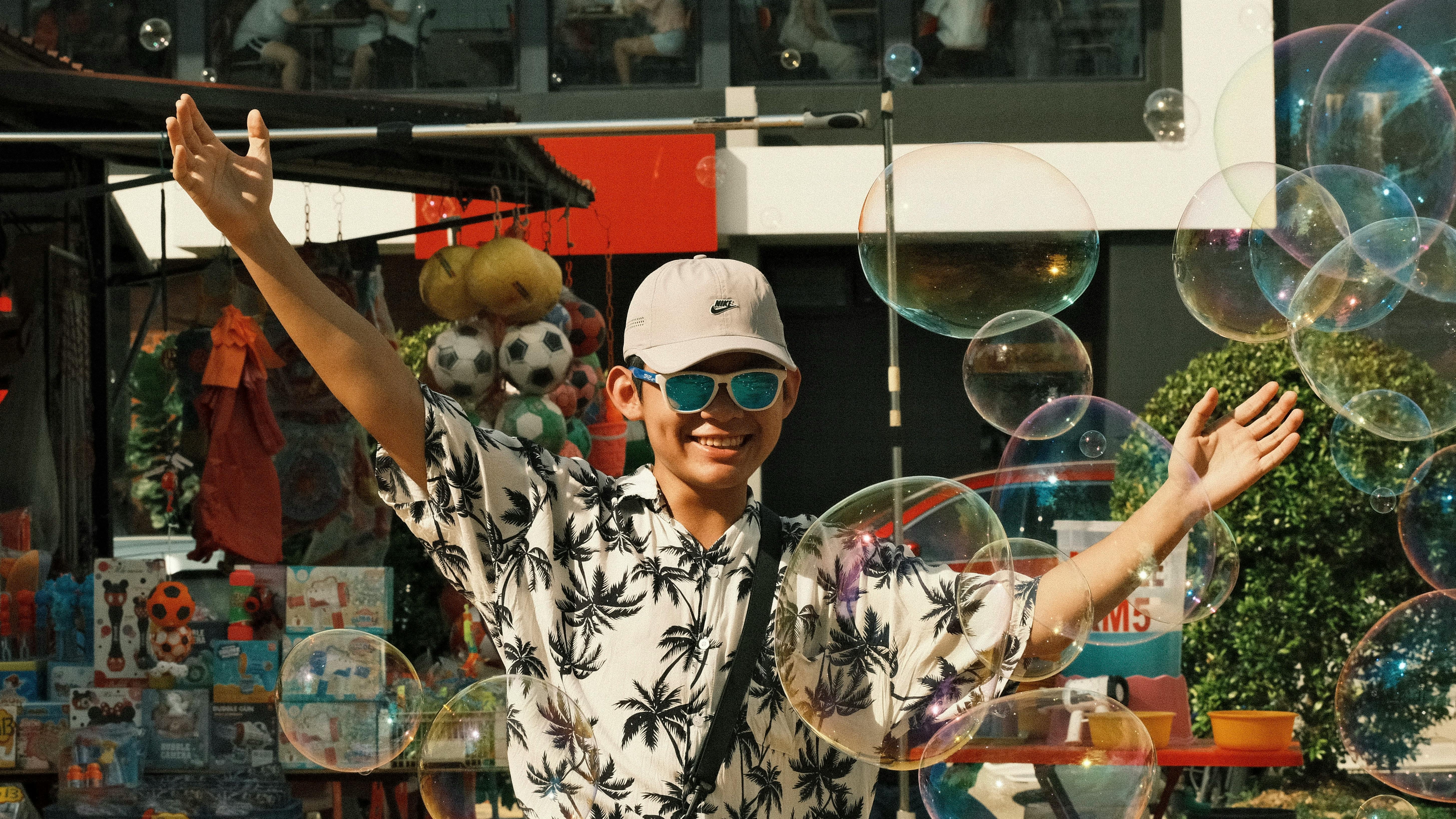 A cheerful man enjoys soap bubbles at an outdoor market stall, wearing sunglasses and a cap.