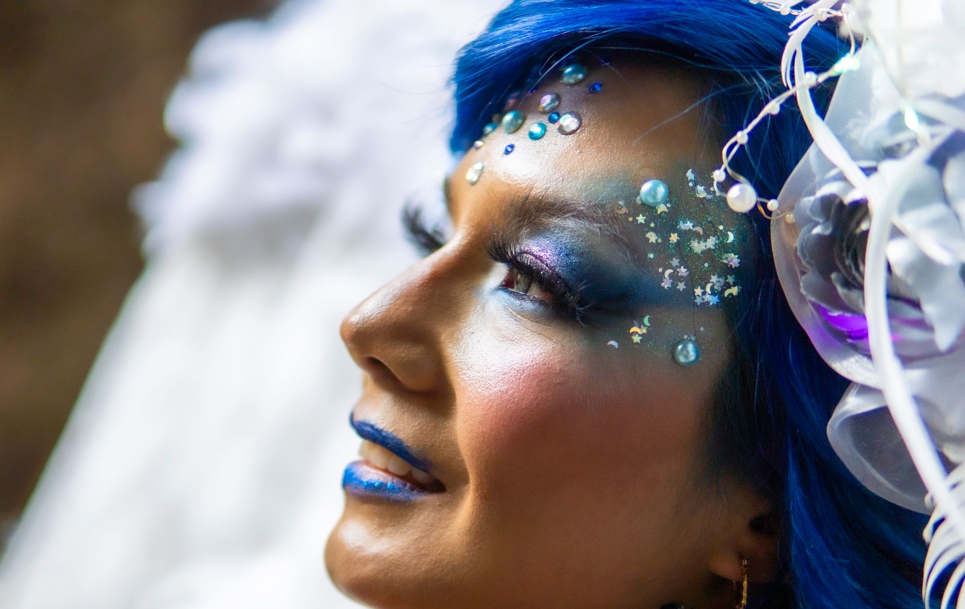 Close-up portrait of a woman in cosplay with blue hair, colorful makeup, and creative costume details.