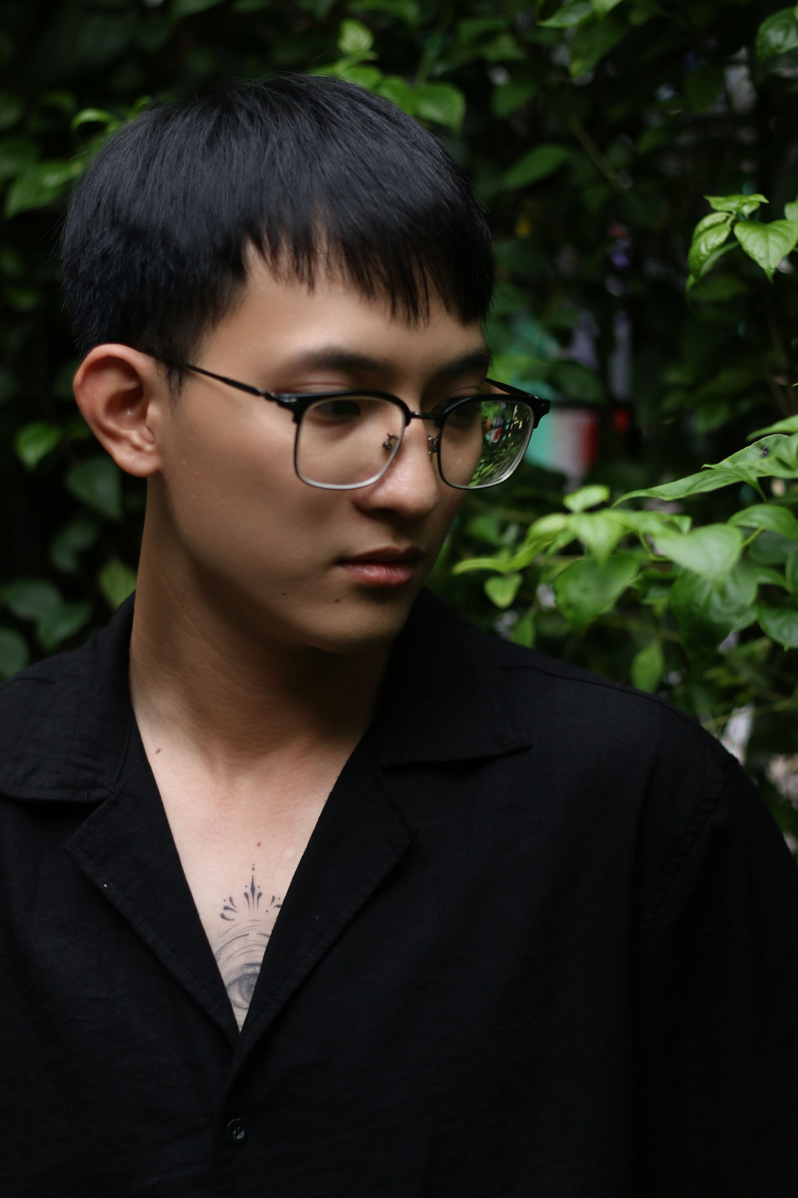 Portrait of a young man standing outdoors among green leaves wearing eyeglasses and a black shirt.