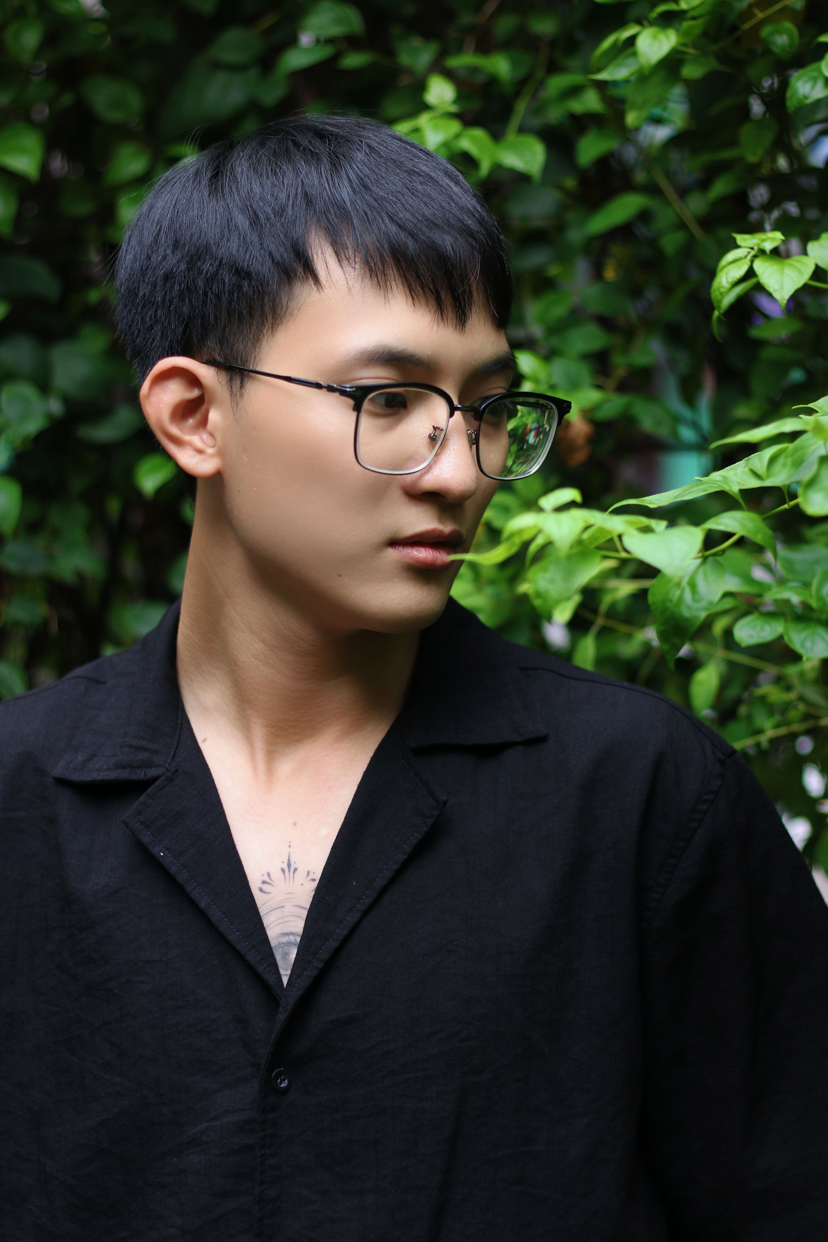 Portrait of a young man wearing glasses, standing among lush green leaves.