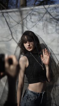 A fashionable woman poses outdoors behind a tulle veil with natural lighting casting shadows.