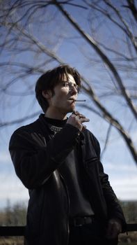 A man in black leather jacket smokes under bare trees in winter sunlight outdoors.