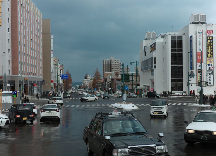 A busy winter street intersection in Otaru, Japan with cars and pedestrians on a cloudy day.
