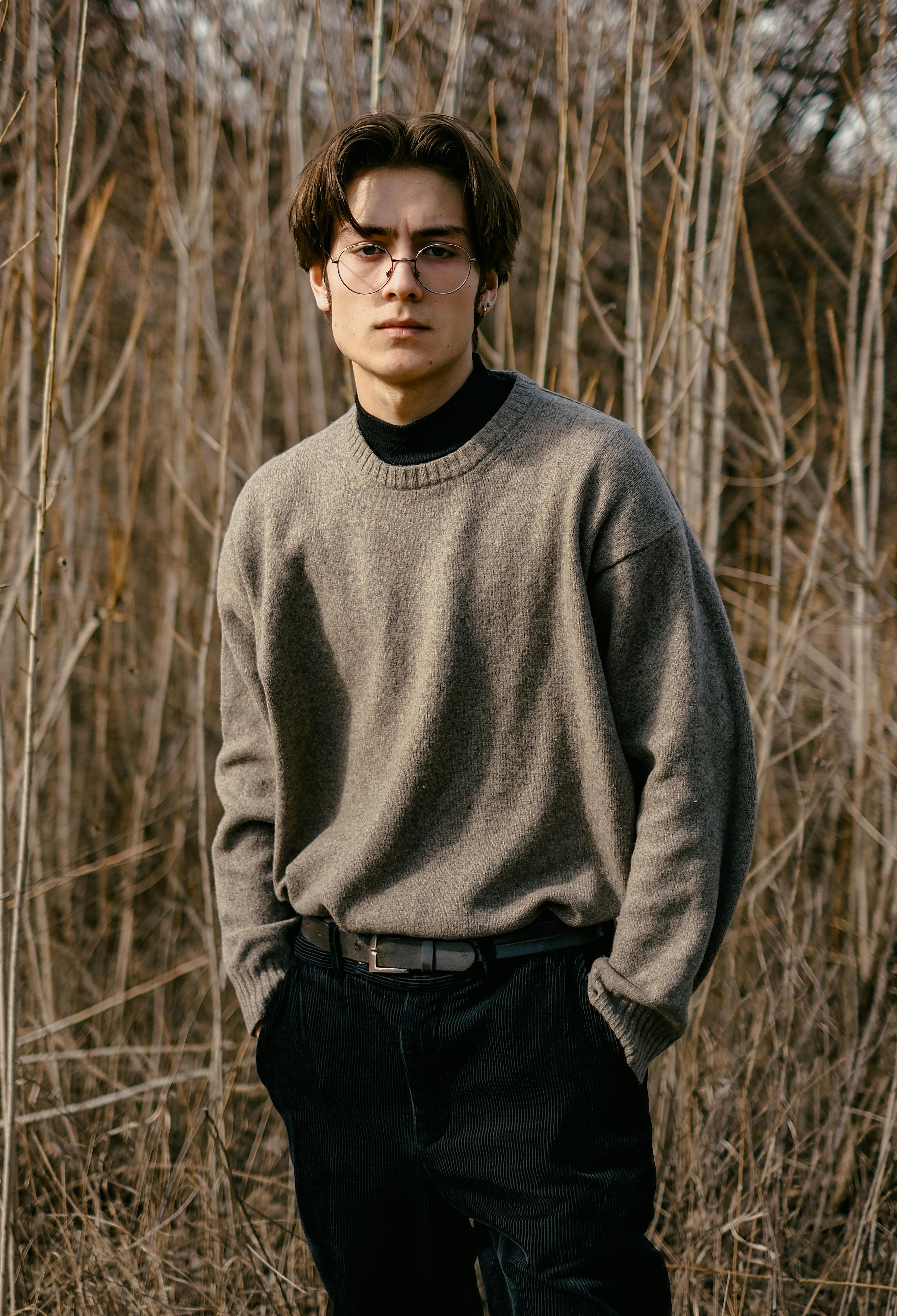 Portrait of a young man in a brown sweater standing among dry branches, embodying autumn fashion.