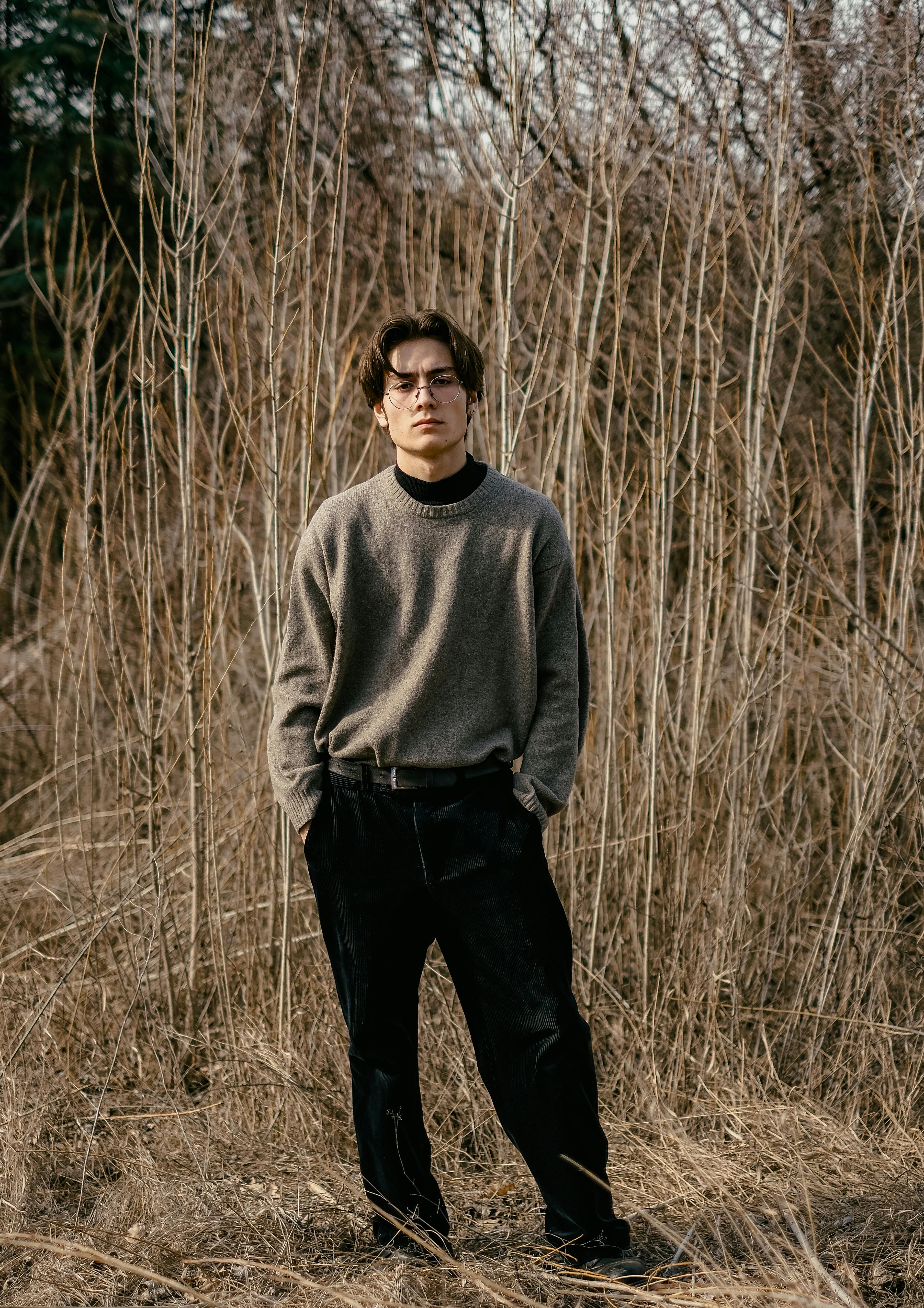 Young man wearing brown sweater and eyeglasses in a fall landscape, hands in pockets.