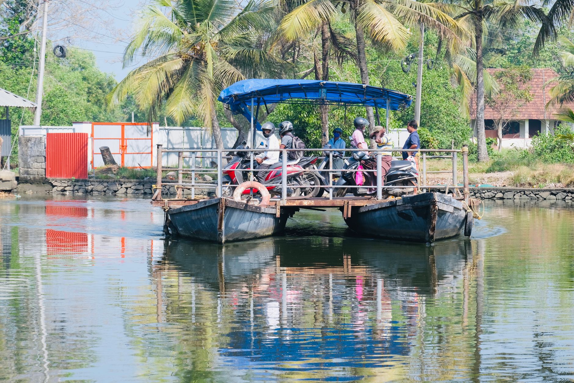 A tropical ferry transports people and motor scooters across a serene lake with lush palm trees in the background.