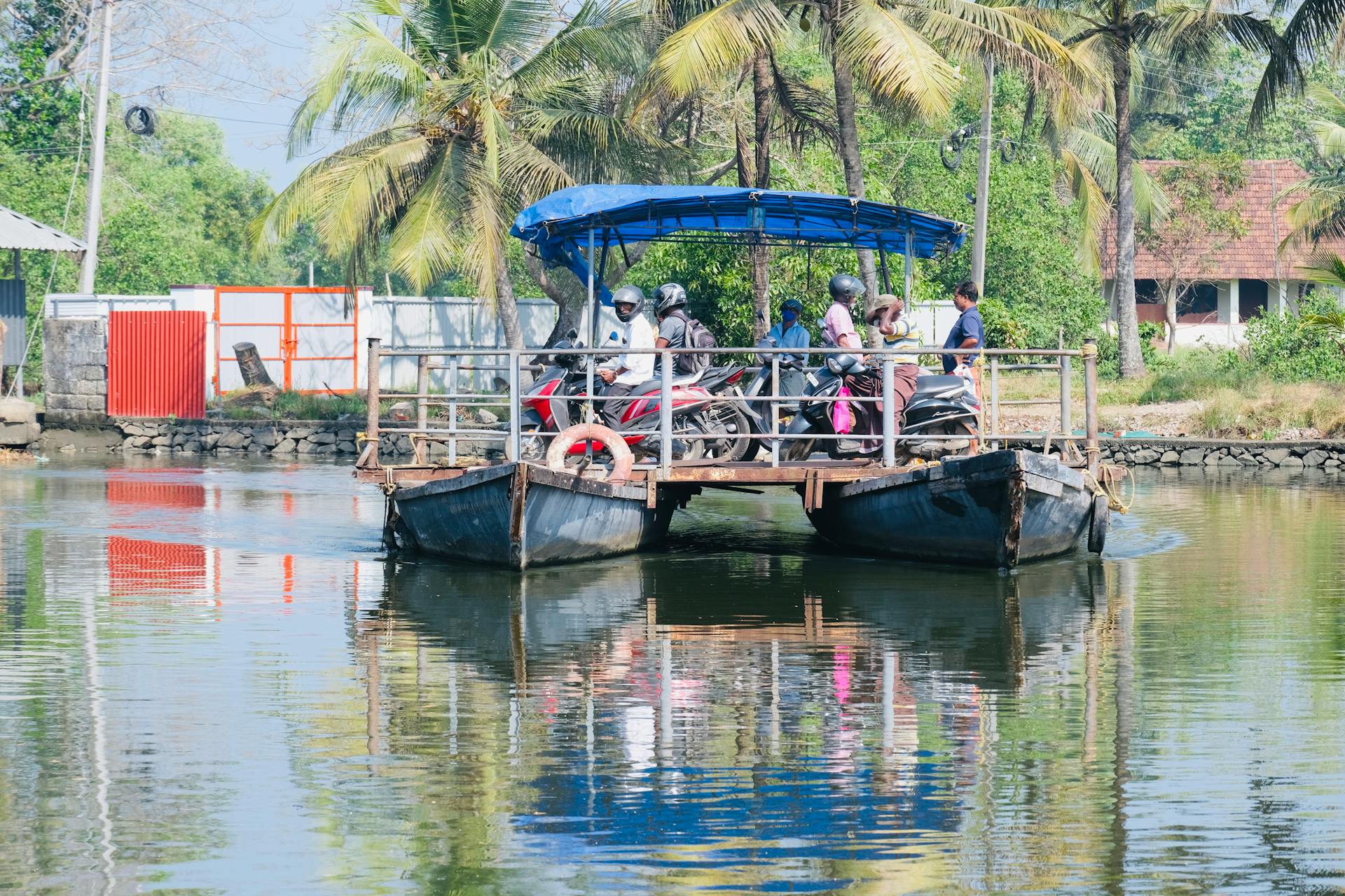 A tropical ferry transports people and motor scooters across a serene lake with lush palm trees in the background.