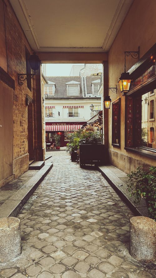 Free A picturesque narrow alleyway in Paris with cobblestone path and vintage lamps leading to a cafe. Stock Photo