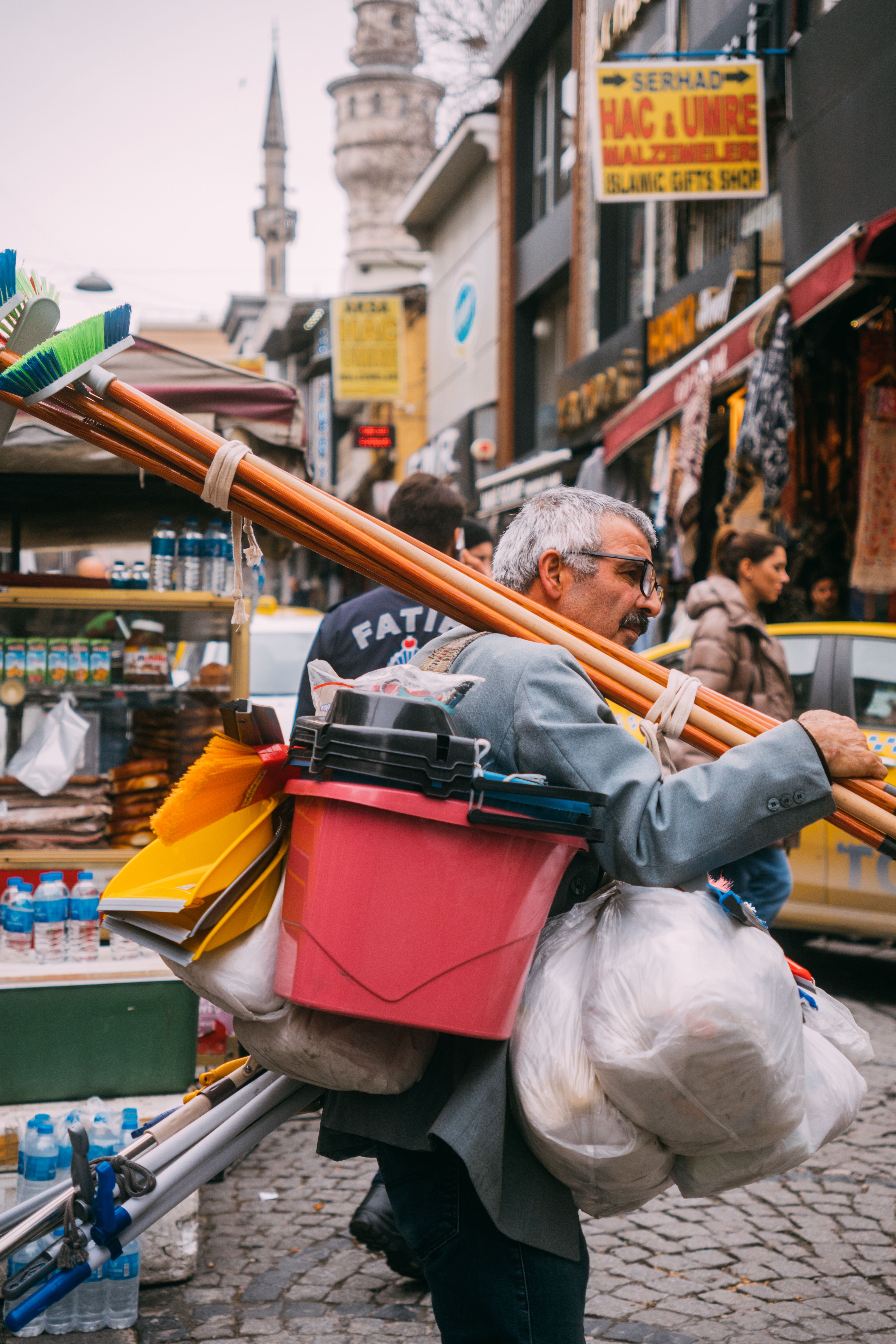 Elderly man carries cleaning tools in vibrant city street market, showcasing urban hustle.