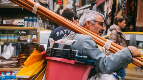 Elderly man carries cleaning tools in vibrant city street market, showcasing urban hustle.