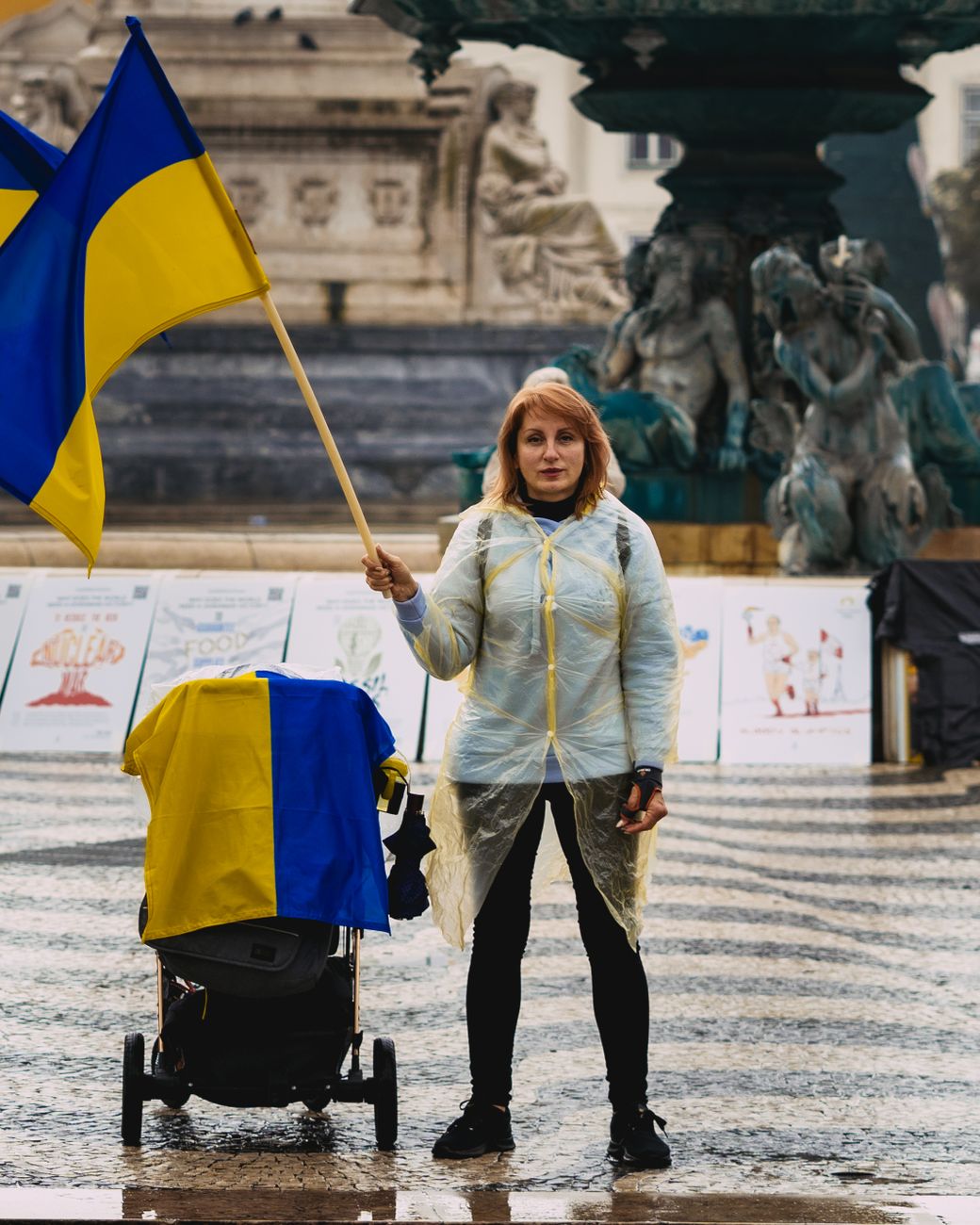 Een vrouw in een regenjas houdt een Oekraïense vlag vast en protesteert in een stedelijke omgeving, terwijl ze naast een standbeeld staat.