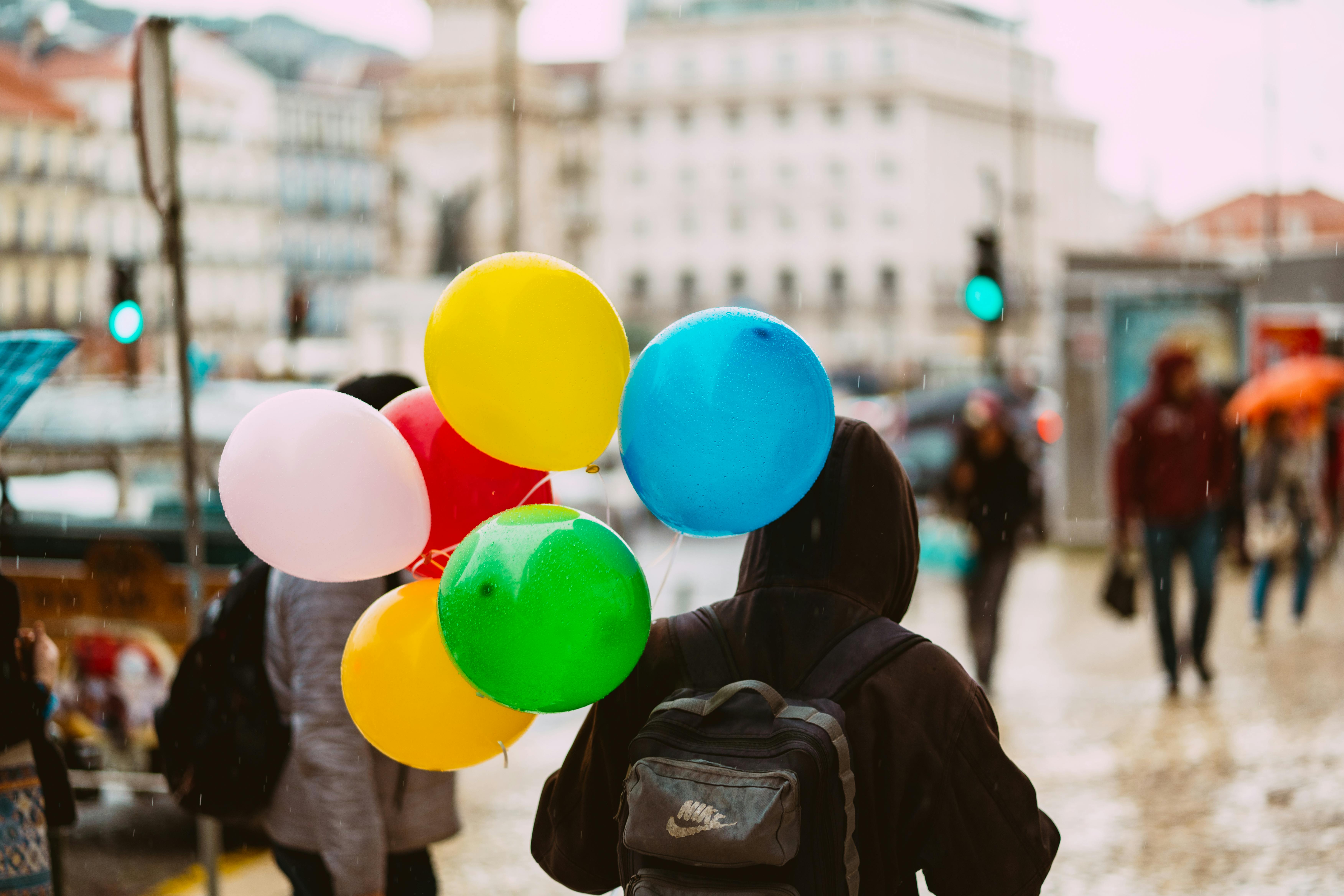 A person in a hoodie holds colorful balloons on a rainy urban street.