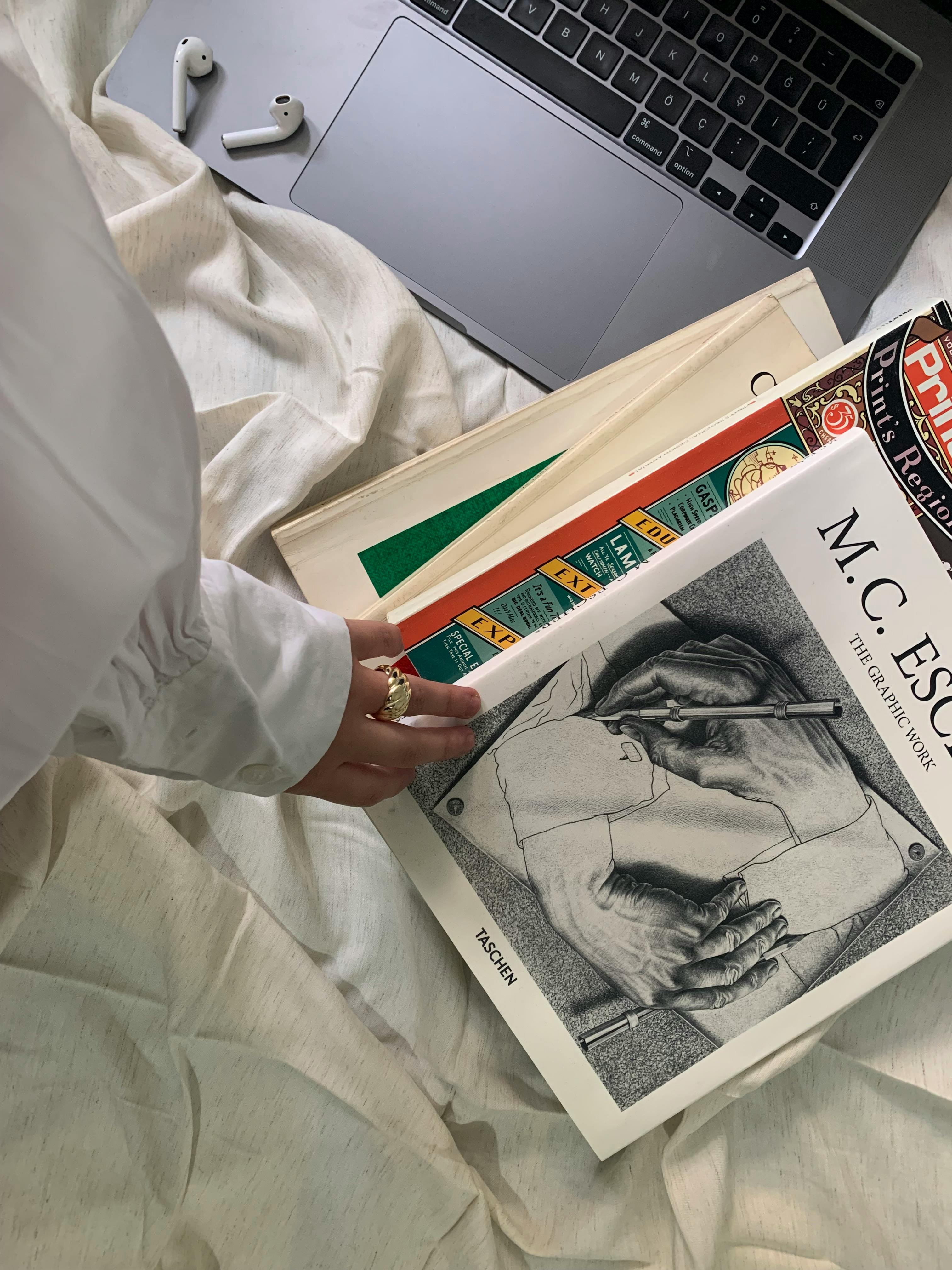 A person reaches for a book beside a laptop and AirPods on a cozy bed.