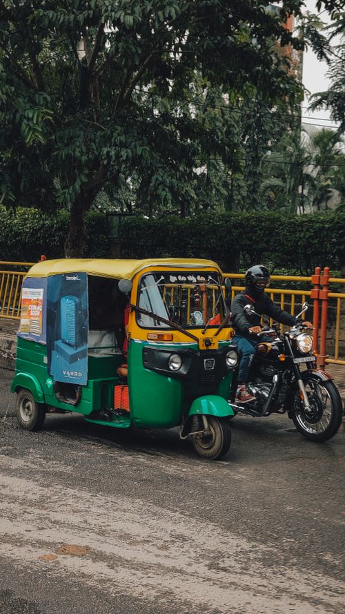 Free Auto rickshaw and motorbike on a rainy city street with lush green trees. Stock Photo