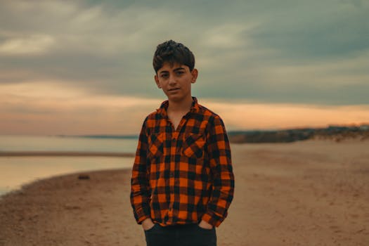 A teenage boy in a checkered shirt standing on the beach during a scenic sunset.