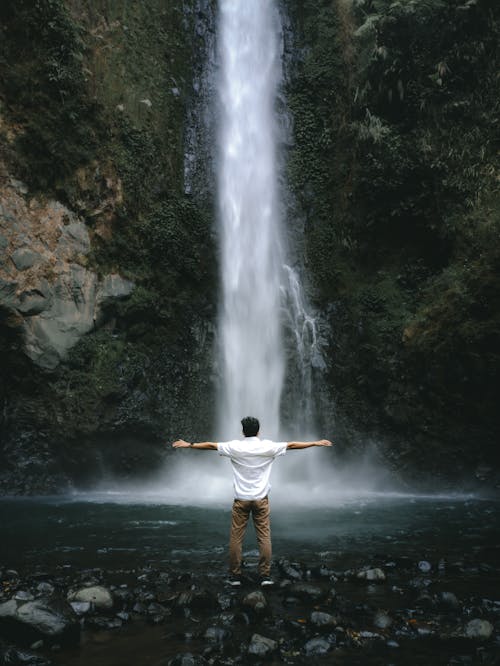 Man in T-shirt Standing with Arms Stretched by Waterfall in Forest