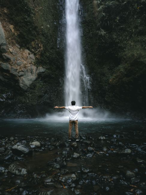 A man stands with arms outstretched facing a grand waterfall, evoking freedom and adventure.