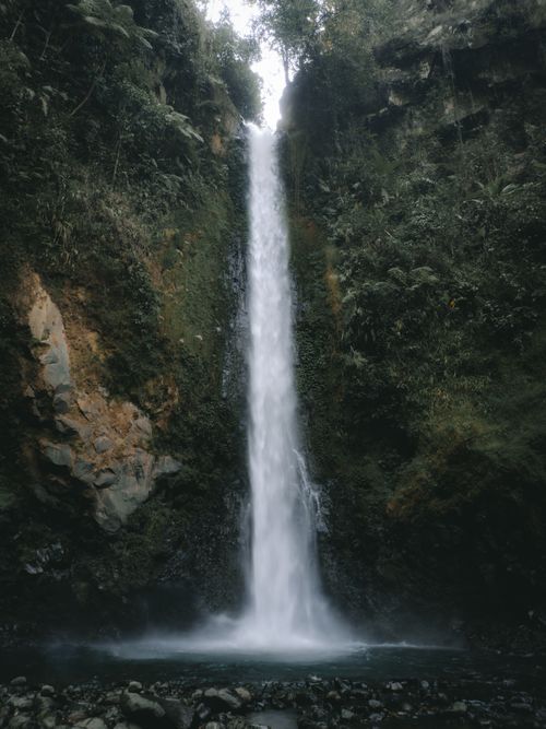 Free A breathtaking waterfall flows through a lush forest, surrounded by rocks and greenery. Stock Photo