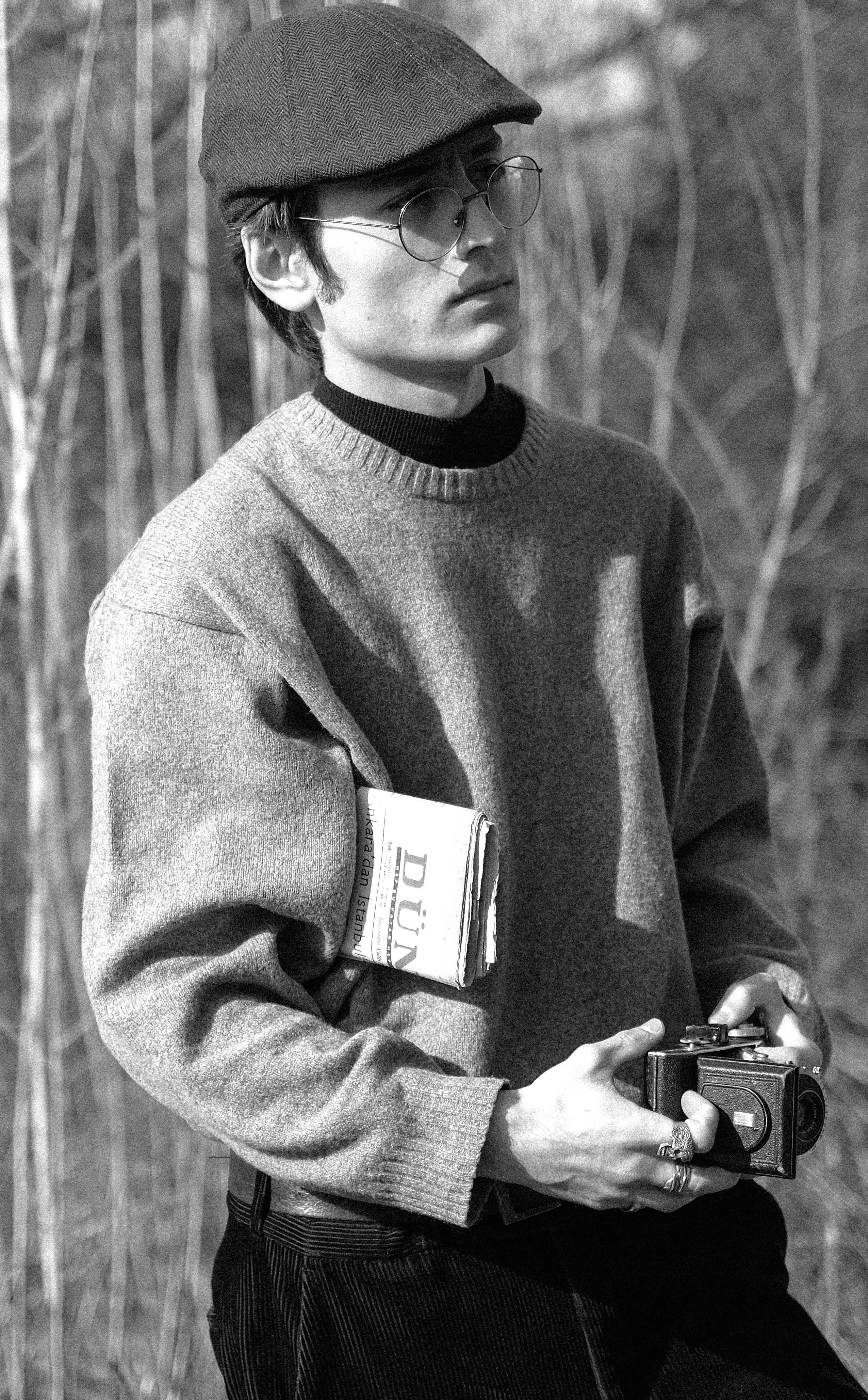 Black and white portrait of a man with eyeglasses and flat cap holding a camera.