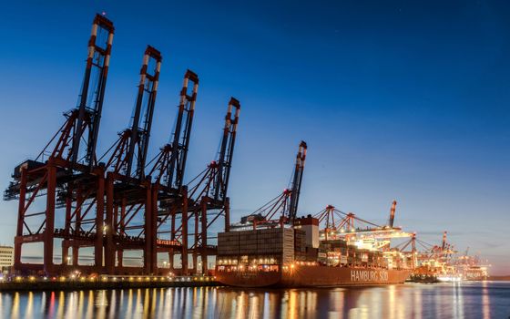 A picturesque evening scene of Hamburg harbor featuring container ships and cranes under a clear sky.