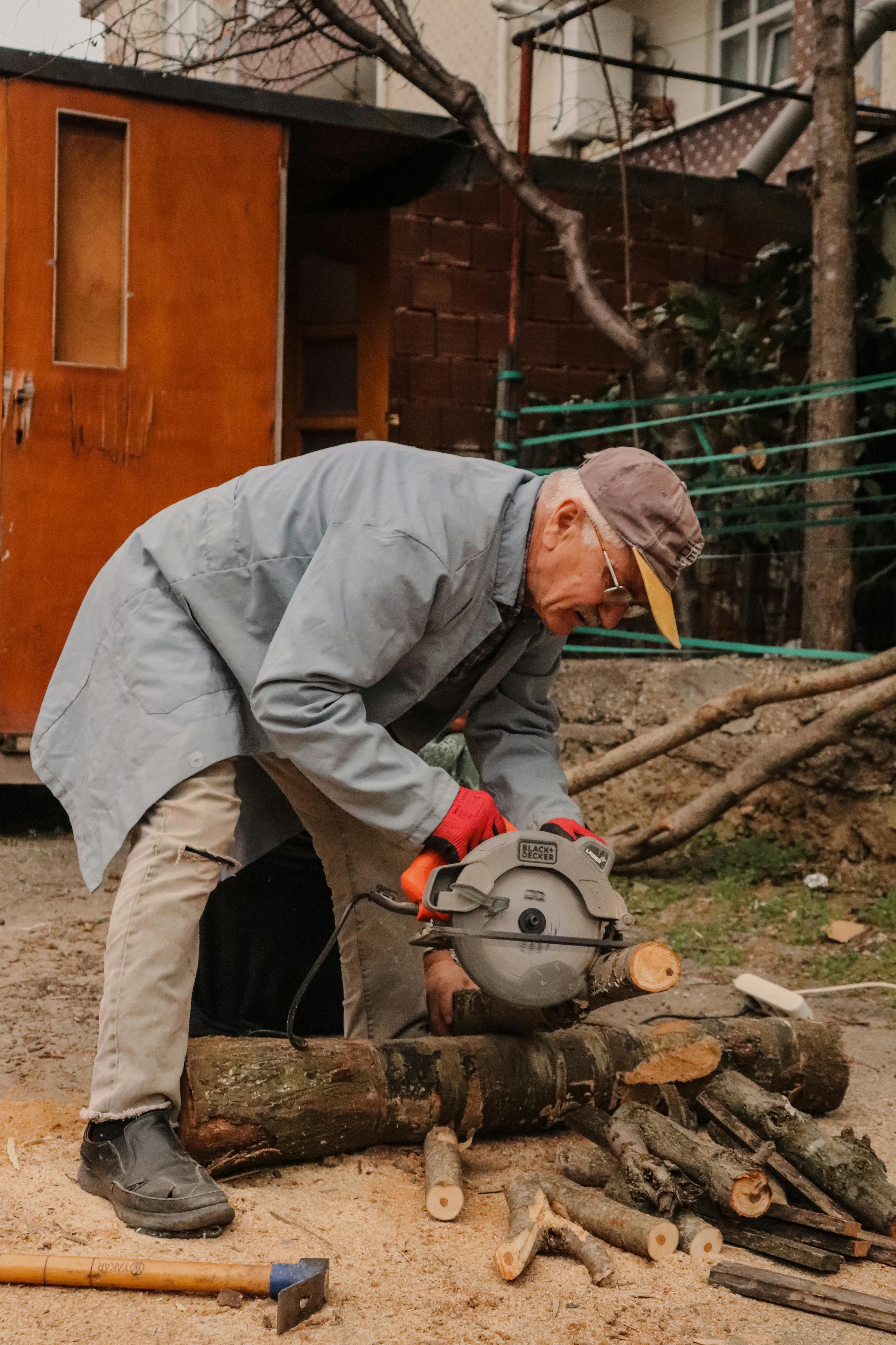 Elderly man cutting logs with circular saw in outdoor yard setting.