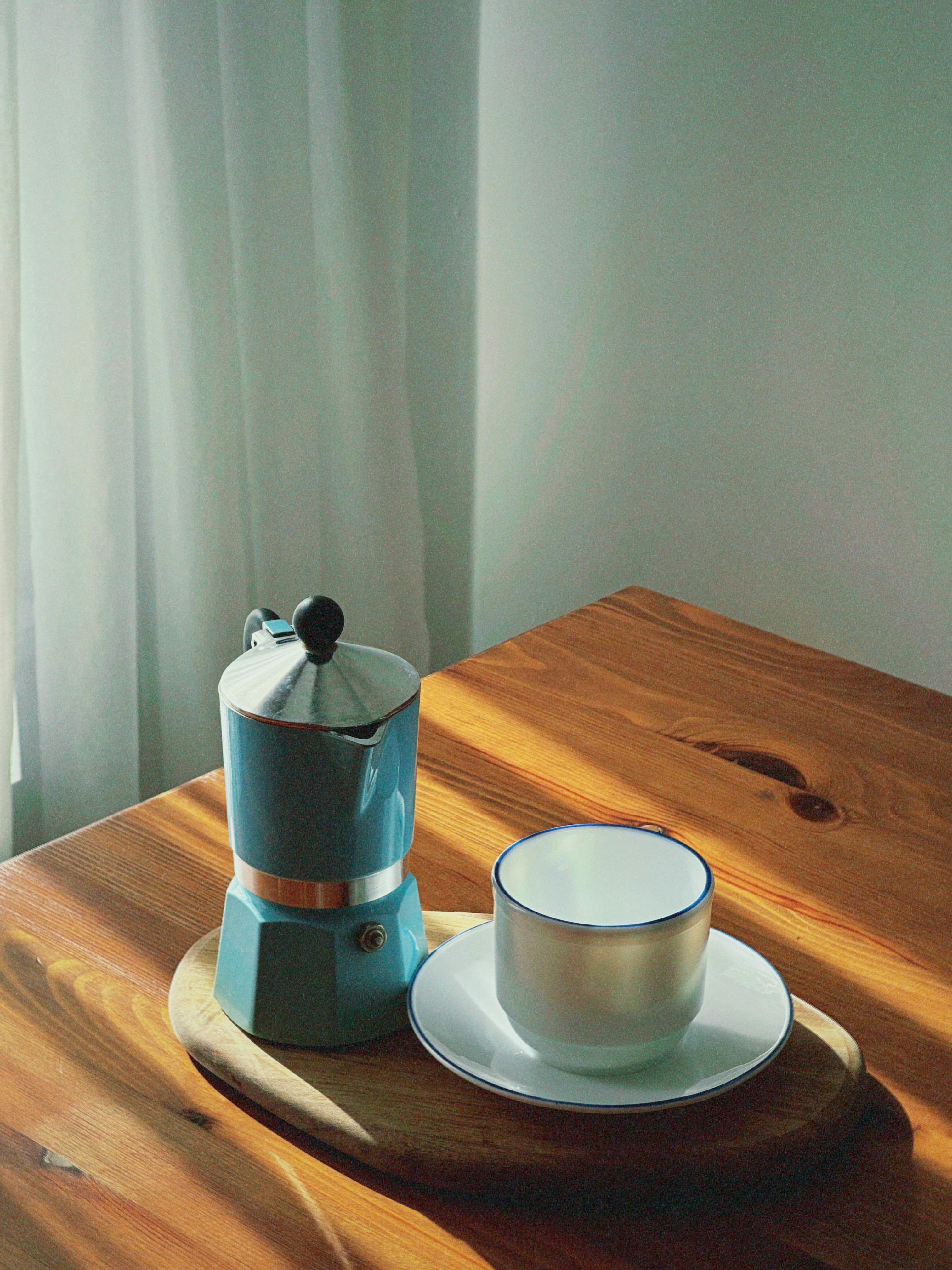 A serene morning scene with a blue Moka pot and coffee cup on a sunlit wooden table.
