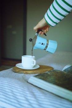 A close-up of a person pouring coffee from a blue moka pot into a white cup indoors.