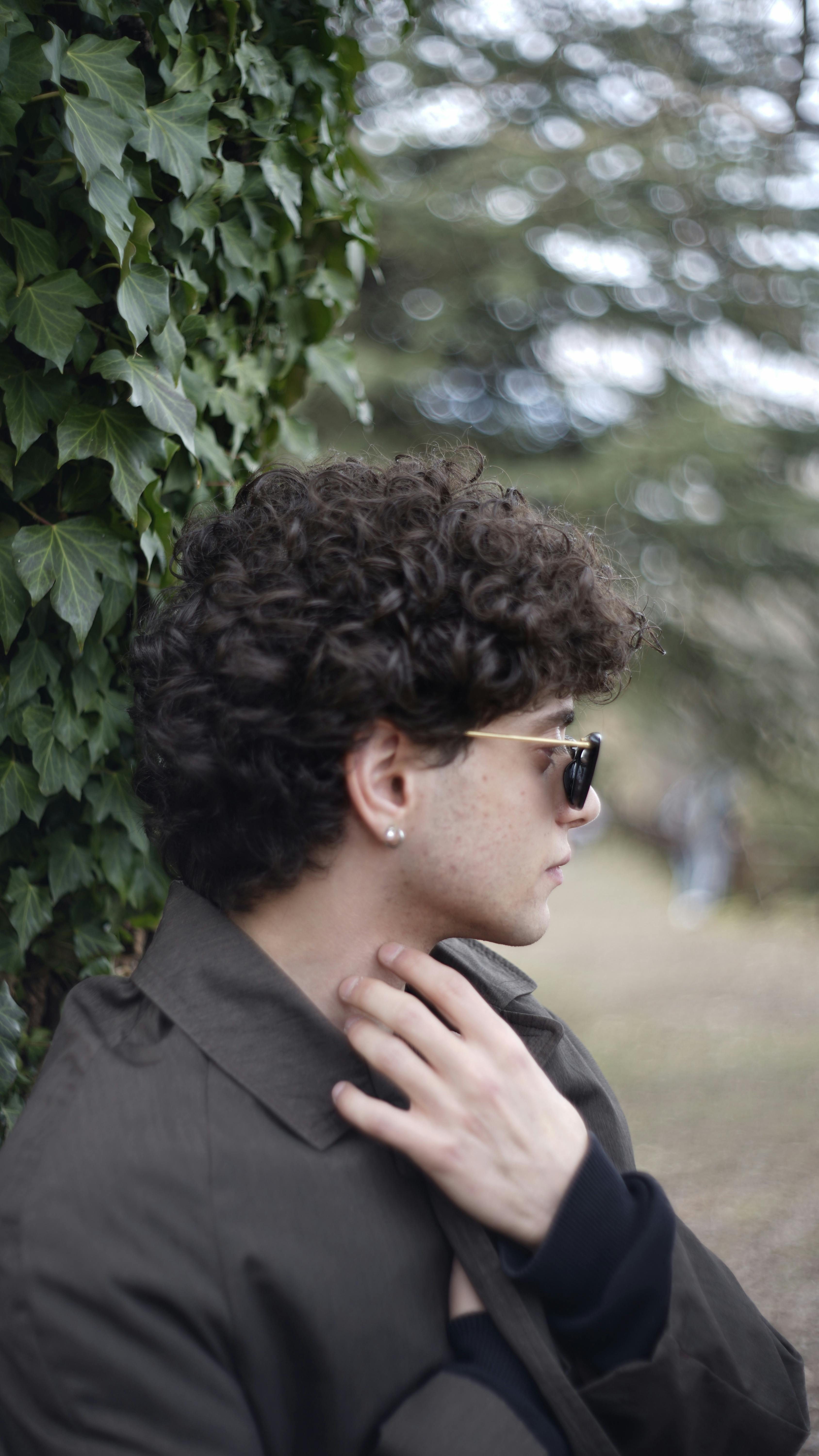 A fashionable young man with sunglasses poses stylishly against a leafy backdrop in an Ankara park.
