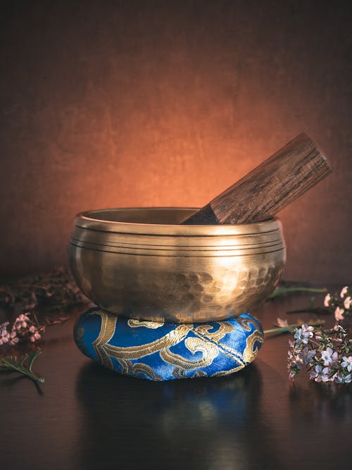 A Tibetan singing bowl sits on a ring cushion amid leaves and flowers, bathed in golden backlight against a dark background