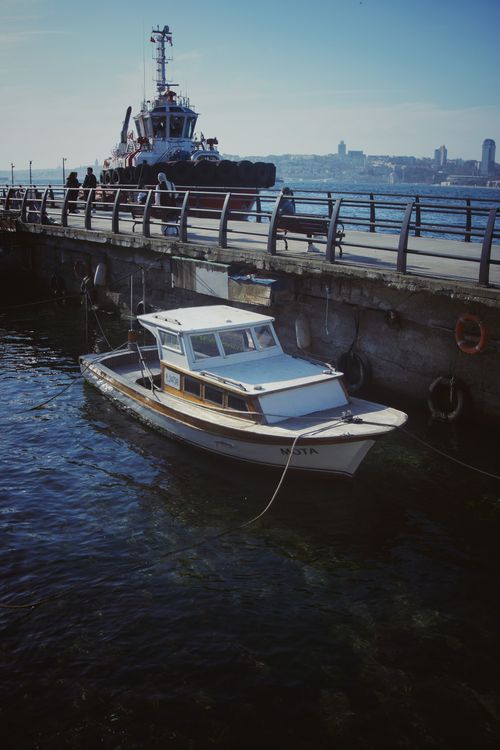 Free A serene harbor scene with a wooden boat moored at a pier and a tugboat in the background. Stock Photo