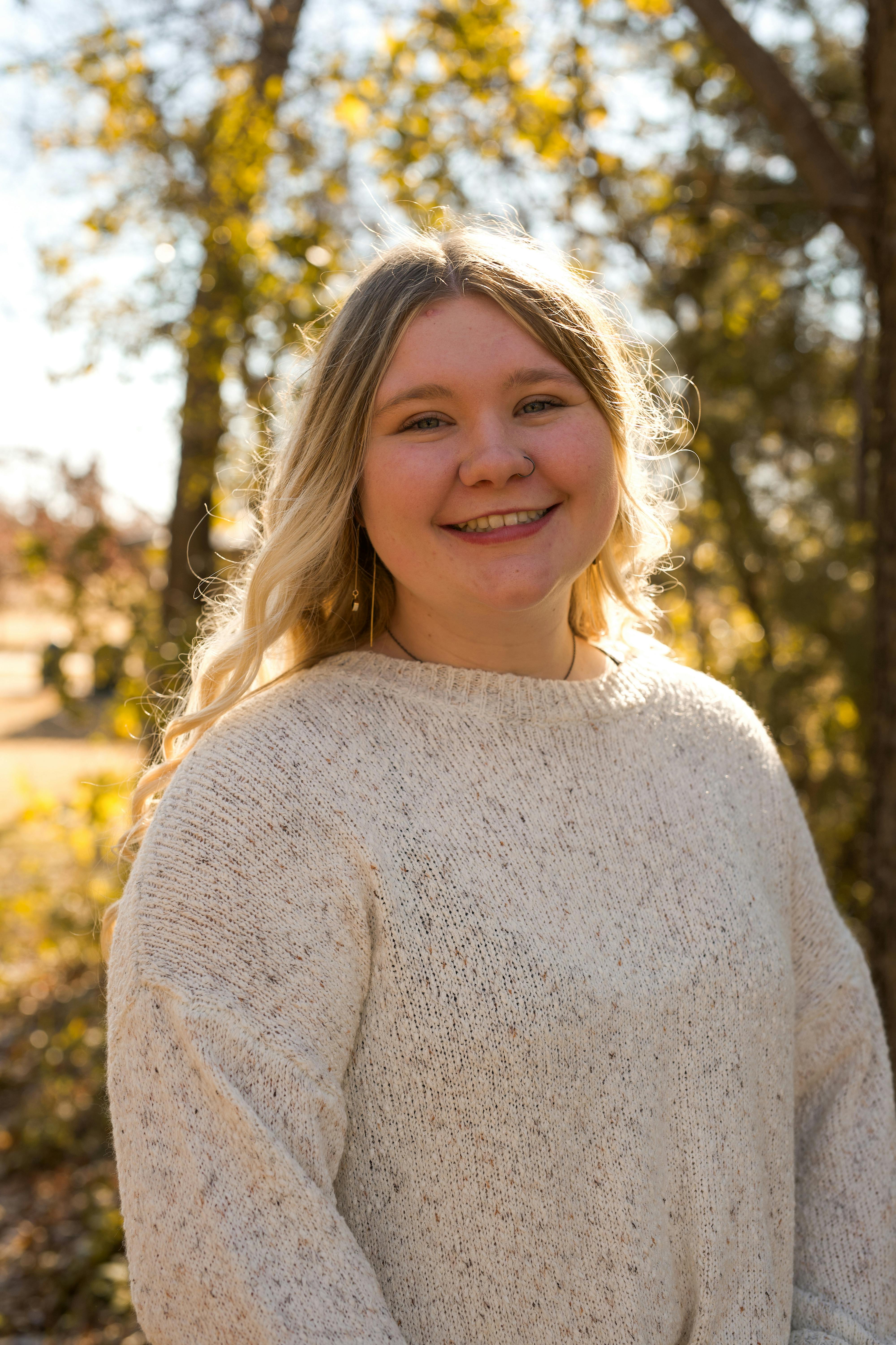 A cheerful young woman poses in a sunlit autumn park wearing a cozy sweater.