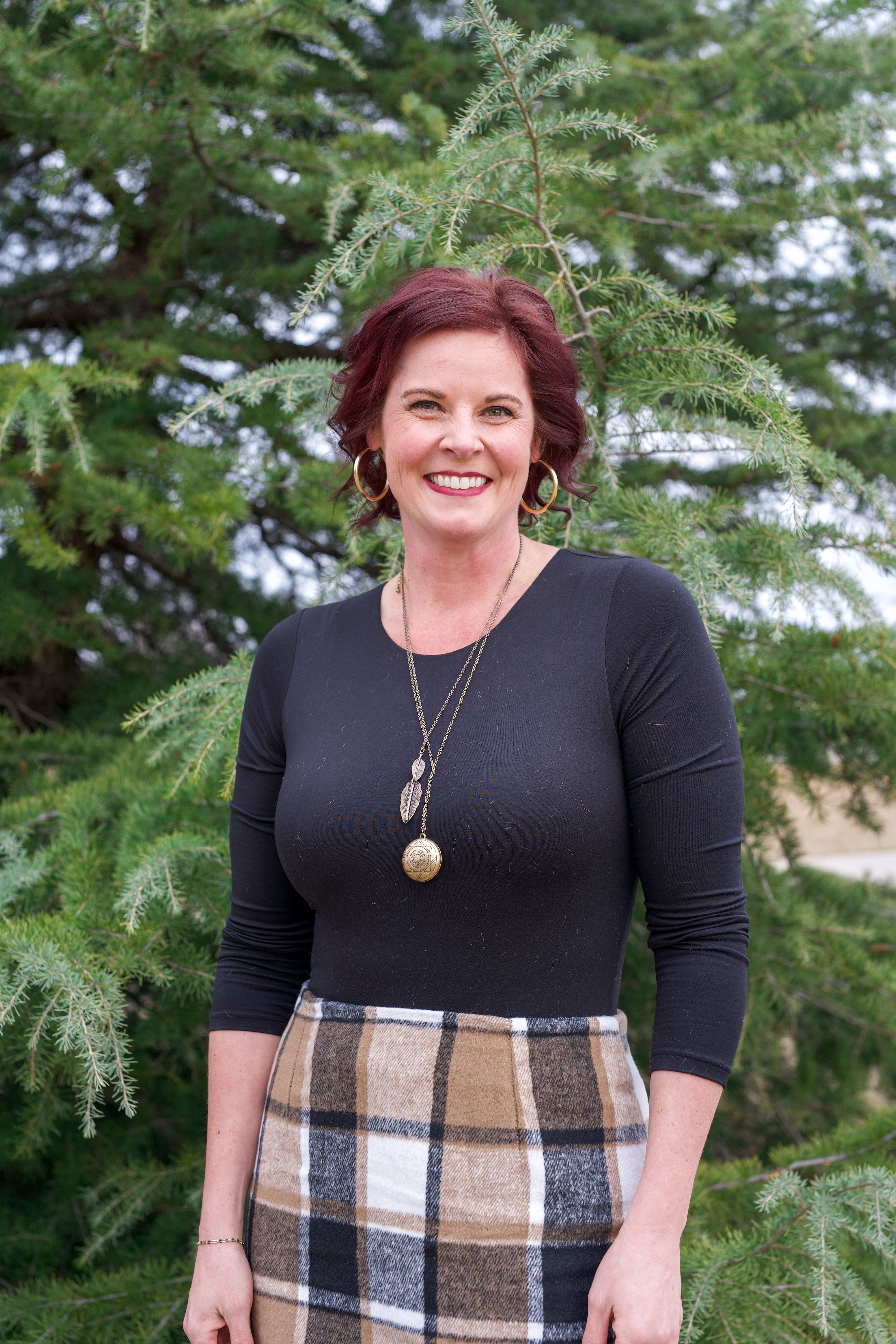 Smiling woman in autumn attire with checked skirt posing outdoors with coniferous trees.