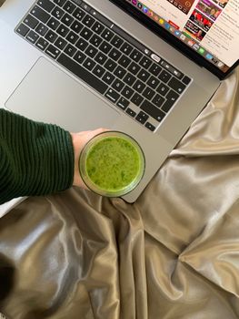 A person holding a green smoothie next to a laptop, suggesting a healthy work-from-home lifestyle.