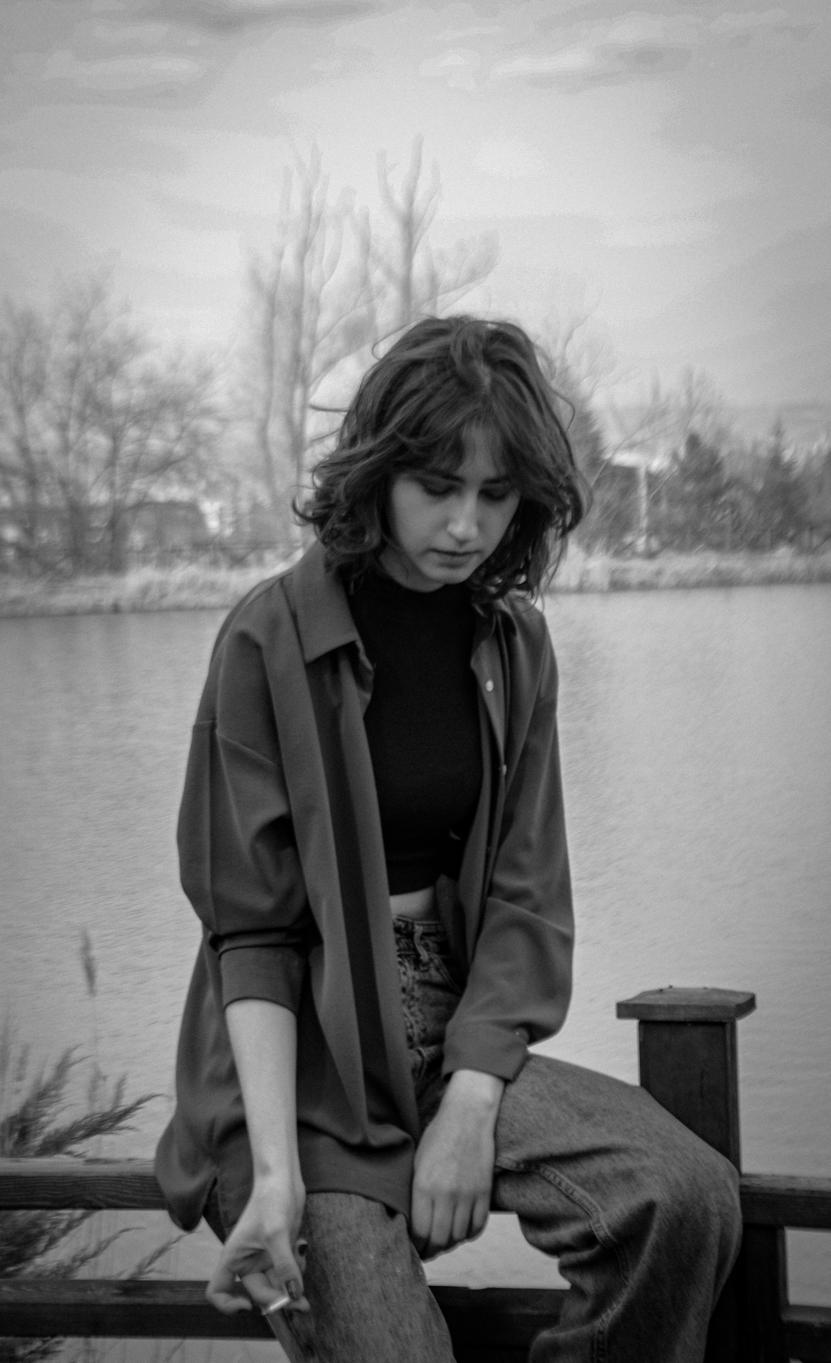 A contemplative woman sitting by the river, holding a cigarette in a black and white setting.