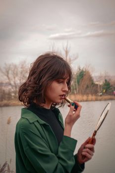 A woman applies lipstick using a handheld mirror near a serene river during the day.