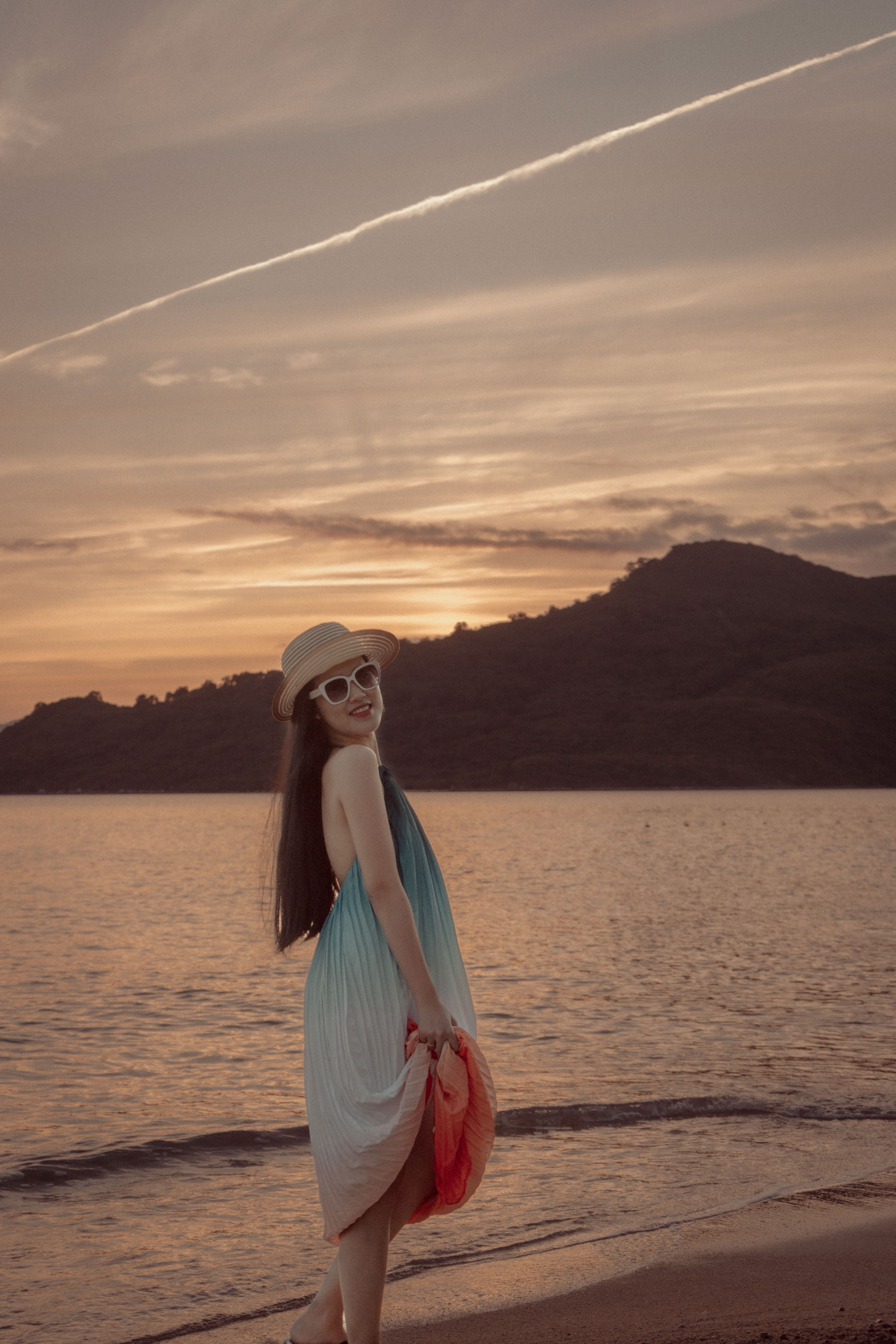 Stylish woman in sundress and sunglasses posing on a sunset beach, capturing a serene vacation vibe.