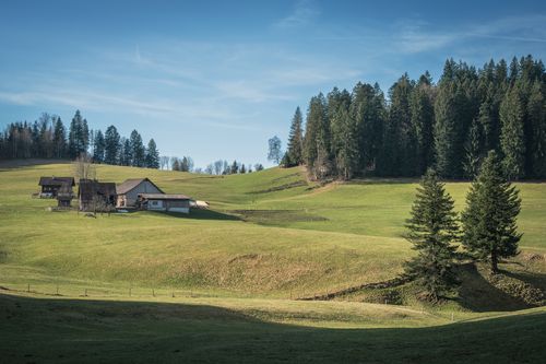 Free Picturesque rural landscape featuring farmhouses, rolling hills, and dense trees under a clear blue sky. Stock Photo