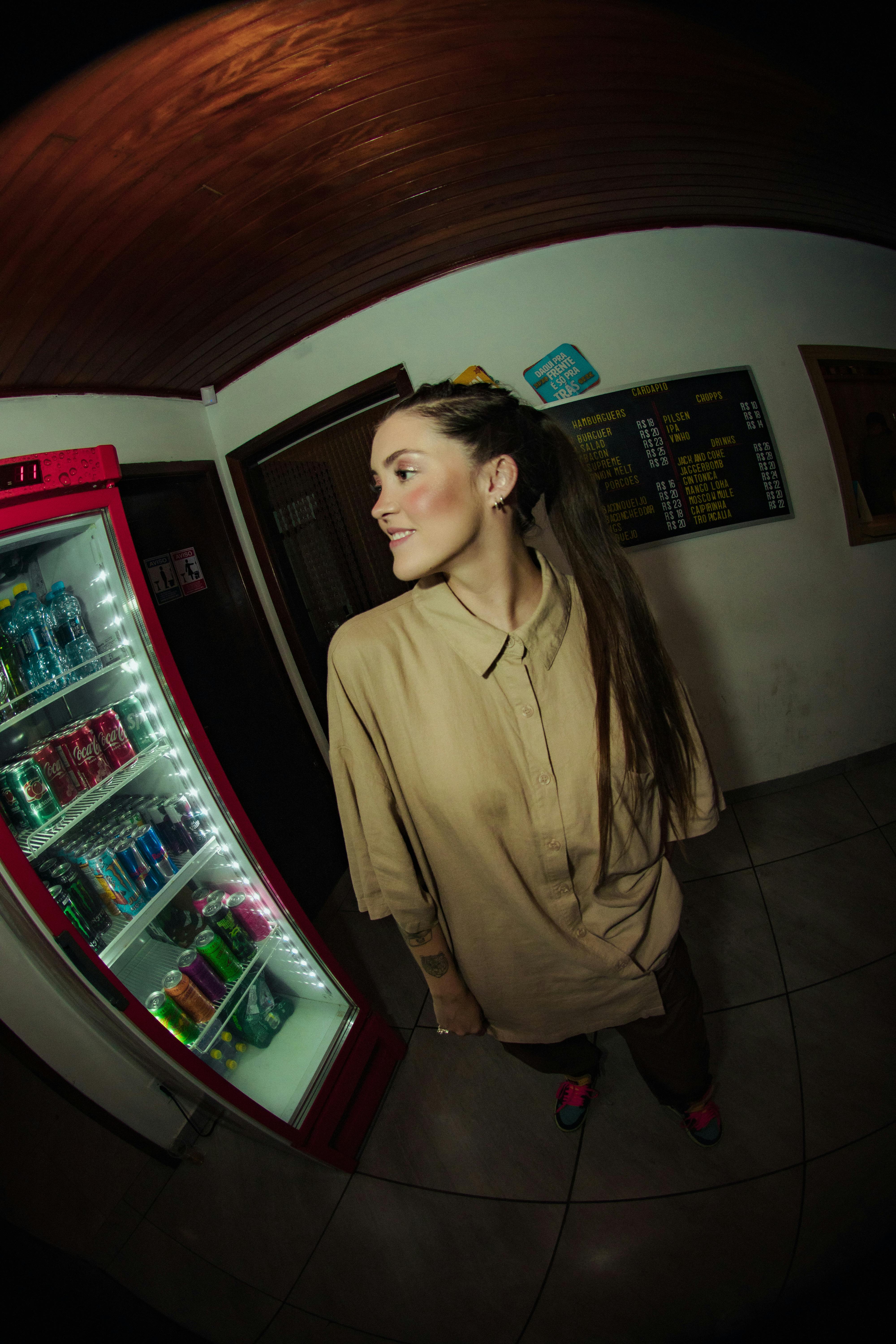 Young woman smiling near a refrigerator in a Brazilian convenience store captured with a fisheye lens.