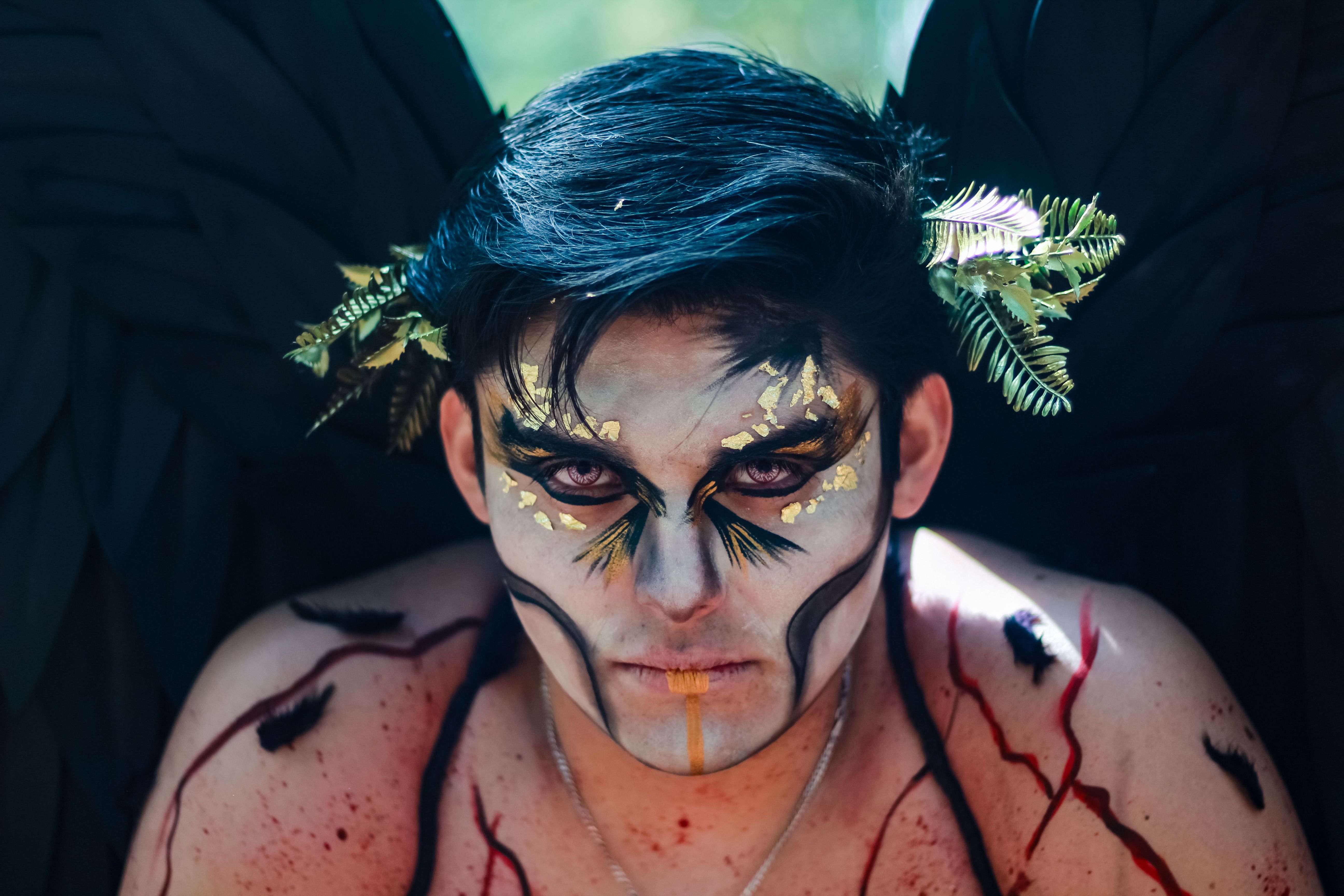 A man with artistic face makeup, black wings, and leaves in a unique fantasy cosplay setup.