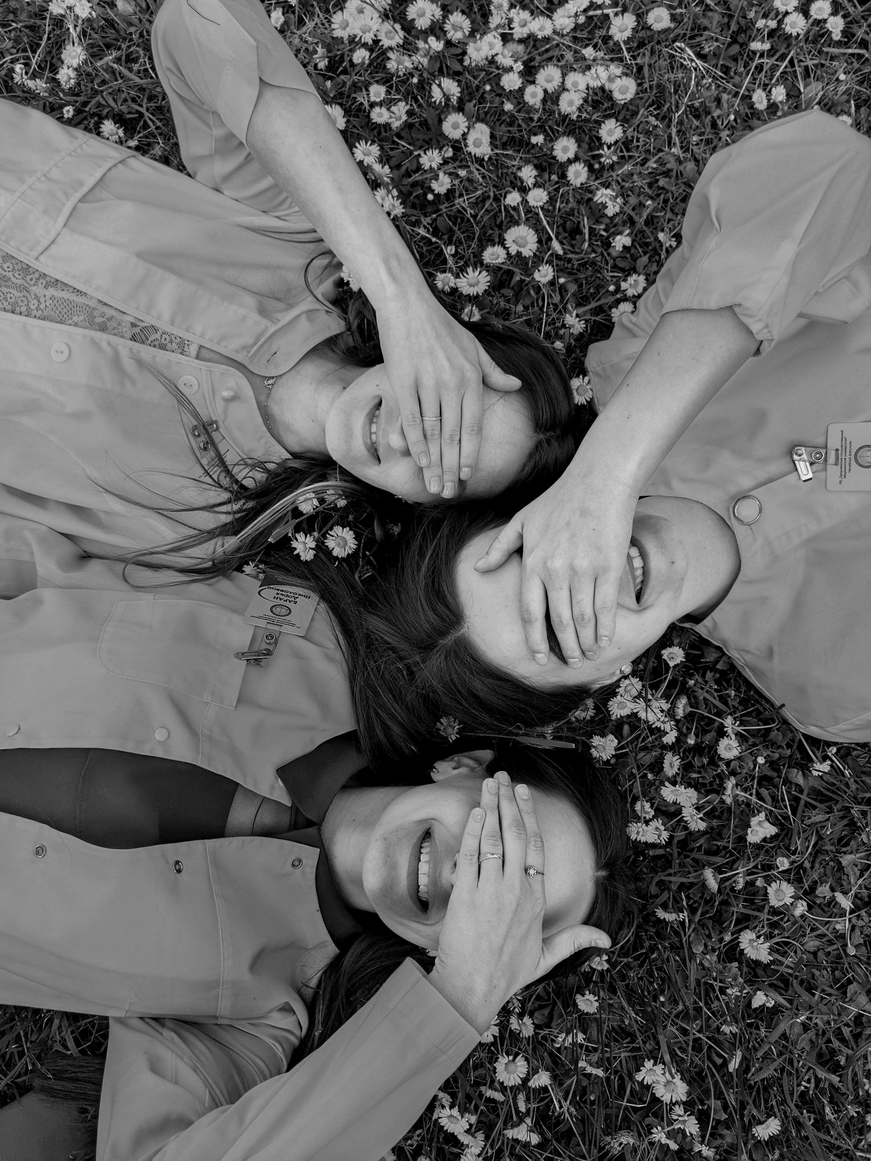 Three women lying on grass, covering eyes while smiling surrounded by flowers.
