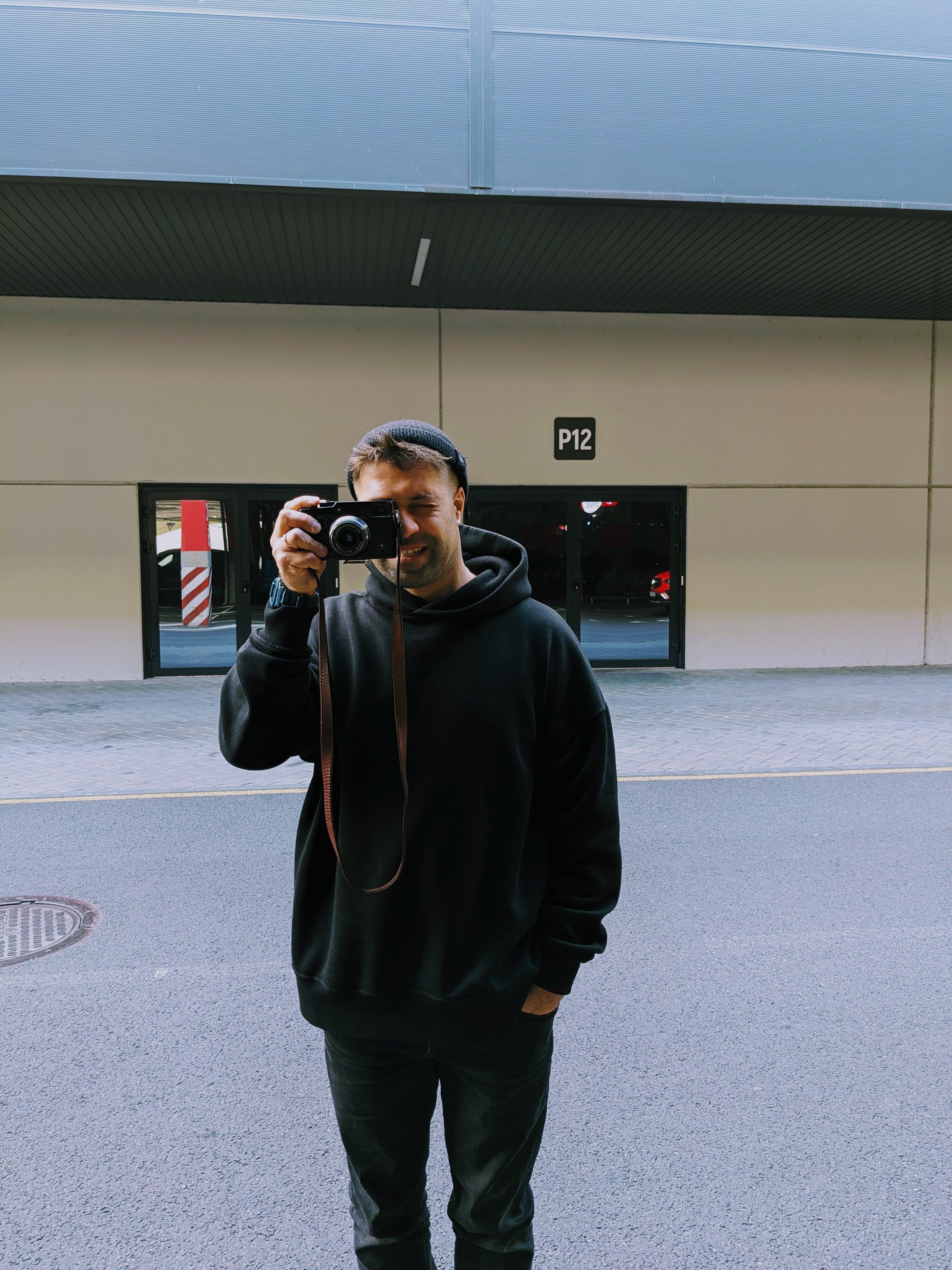 A photographer in a black hoodie takes a picture outdoors near a building marked P12.
