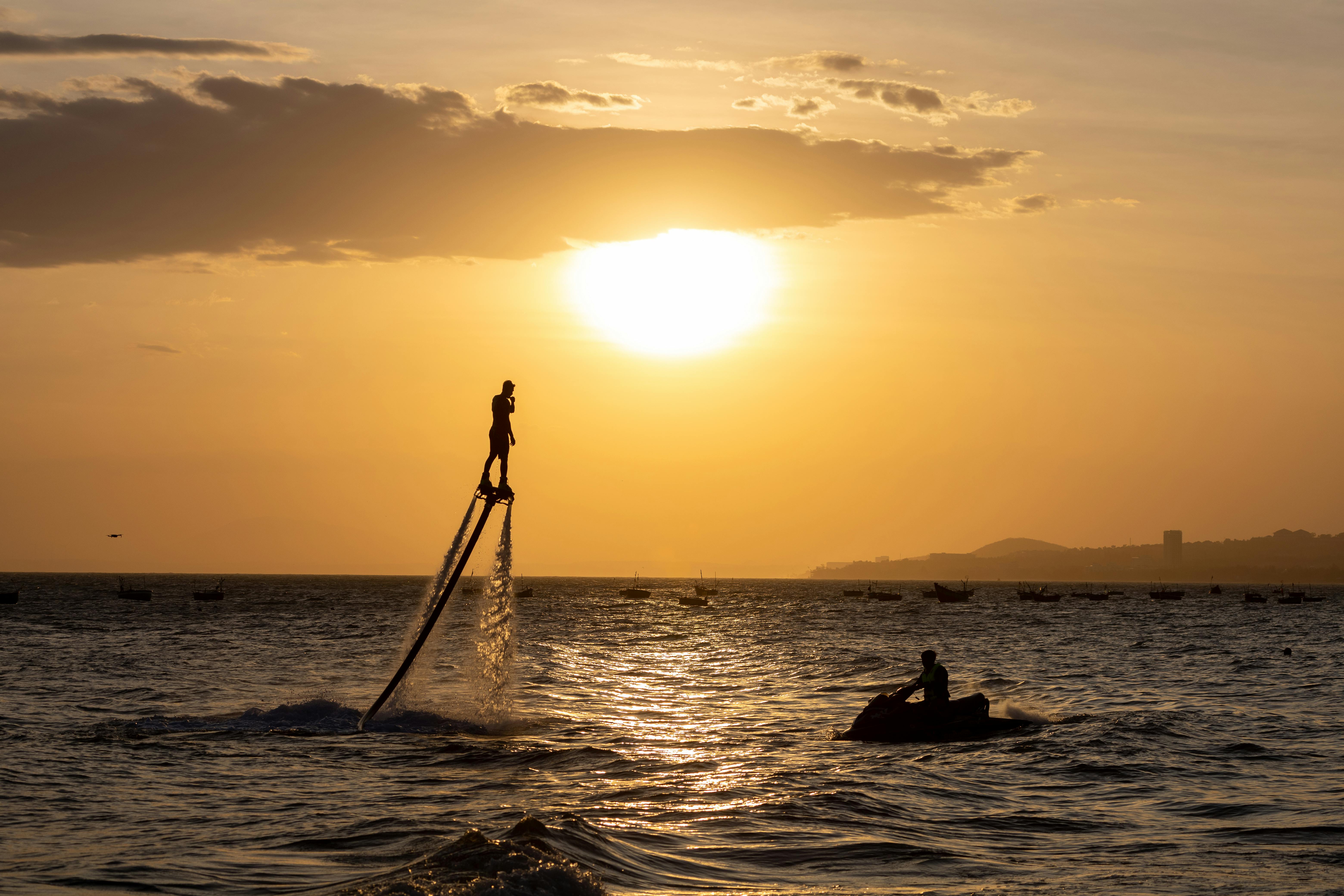 A person flyboarding at sunset over a vibrant sea with a jet ski nearby.